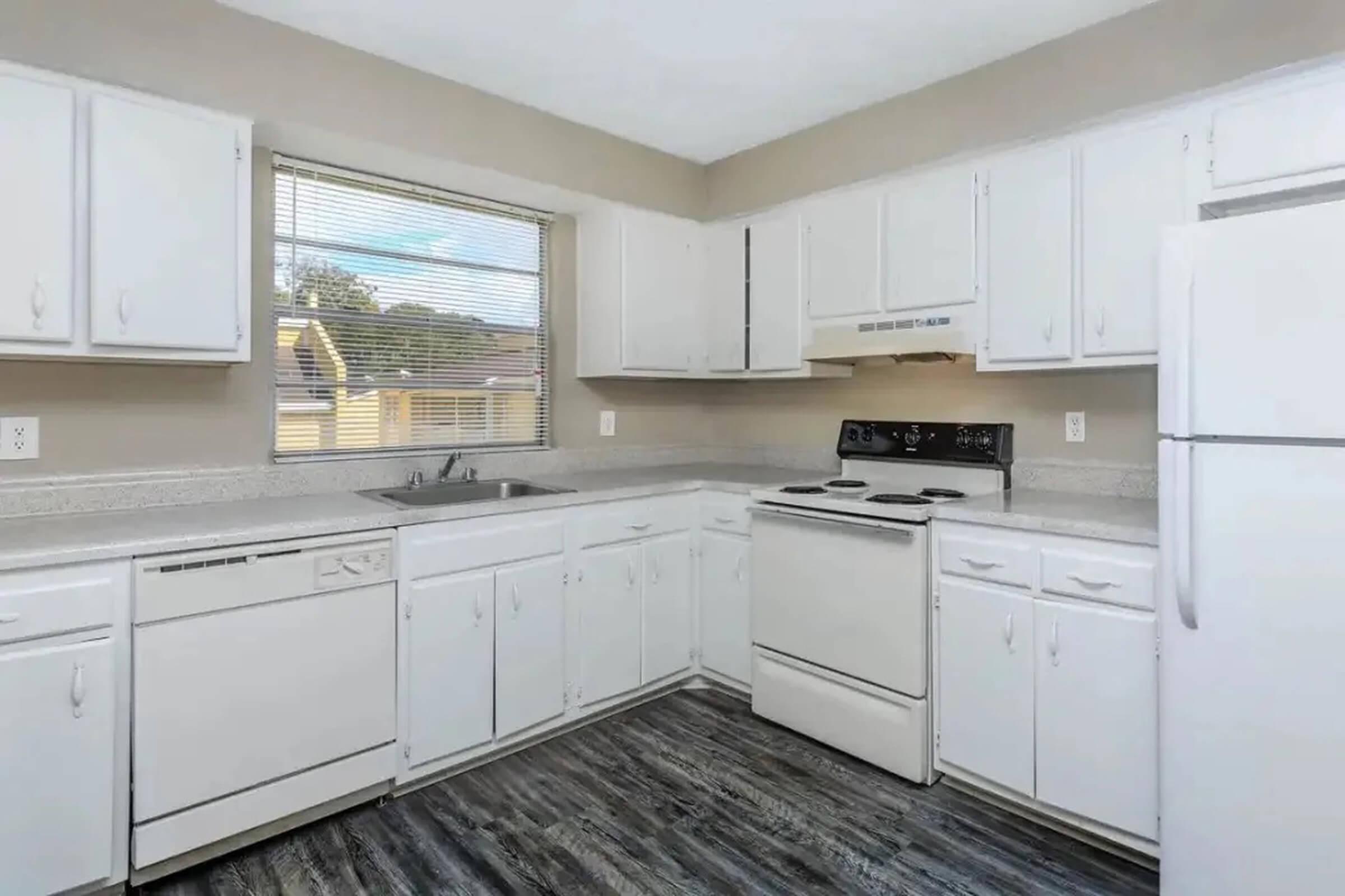 A modern kitchen featuring white cabinetry, a sink below a window, an electric stove, a refrigerator, and a built-in dishwasher. The countertops are light gray, and the flooring is dark, creating a contrast in the space. Bright, natural light fills the room.