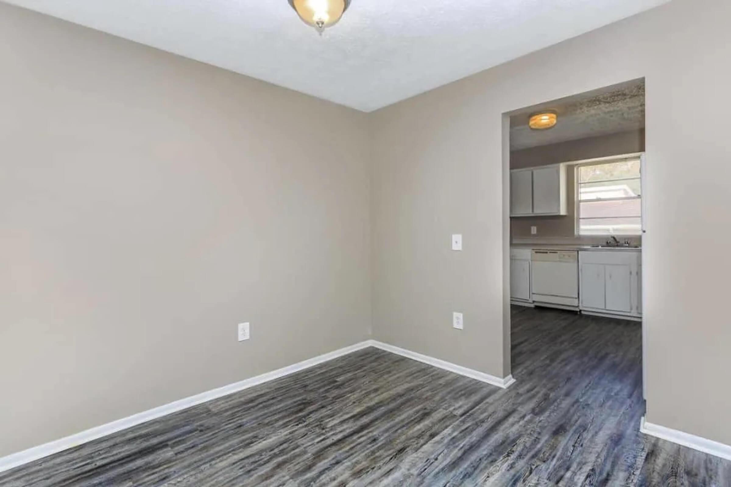 A small, empty room with light beige walls and a wooden floor. There is a doorway leading to a kitchen area with white cabinets visible. A light fixture hangs from the ceiling, providing illumination in the space. The overall ambiance is bright and inviting, suitable for various uses.