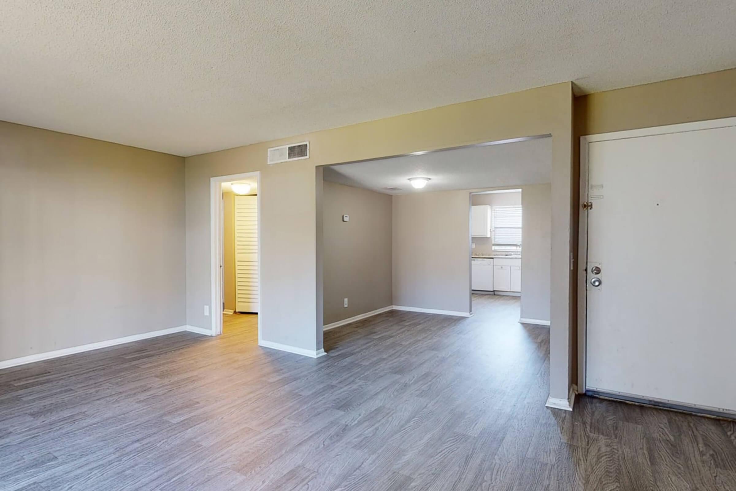 A spacious, empty living room with light-colored walls and laminate flooring. A doorway leads to a small hallway, and another doorway opens to a kitchen with natural light coming through the window. The entrance door is on the right side of the image.