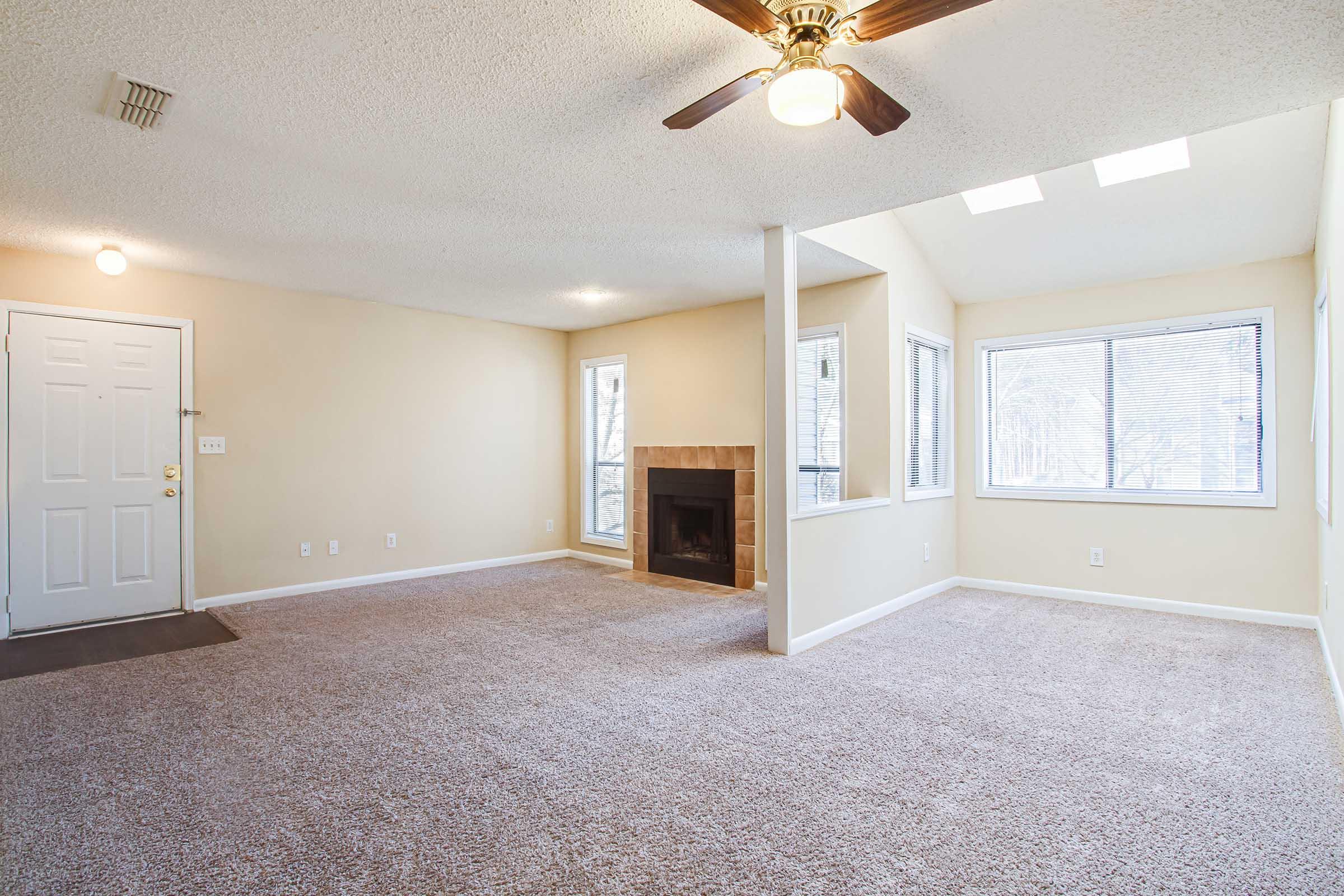 A spacious, well-lit living room featuring neutral-colored walls and carpet. There is a ceiling fan and a fireplace centrally located. Large windows allow natural light, and the open layout connects to other areas of the home. The entrance is visible with a front door on the left side.