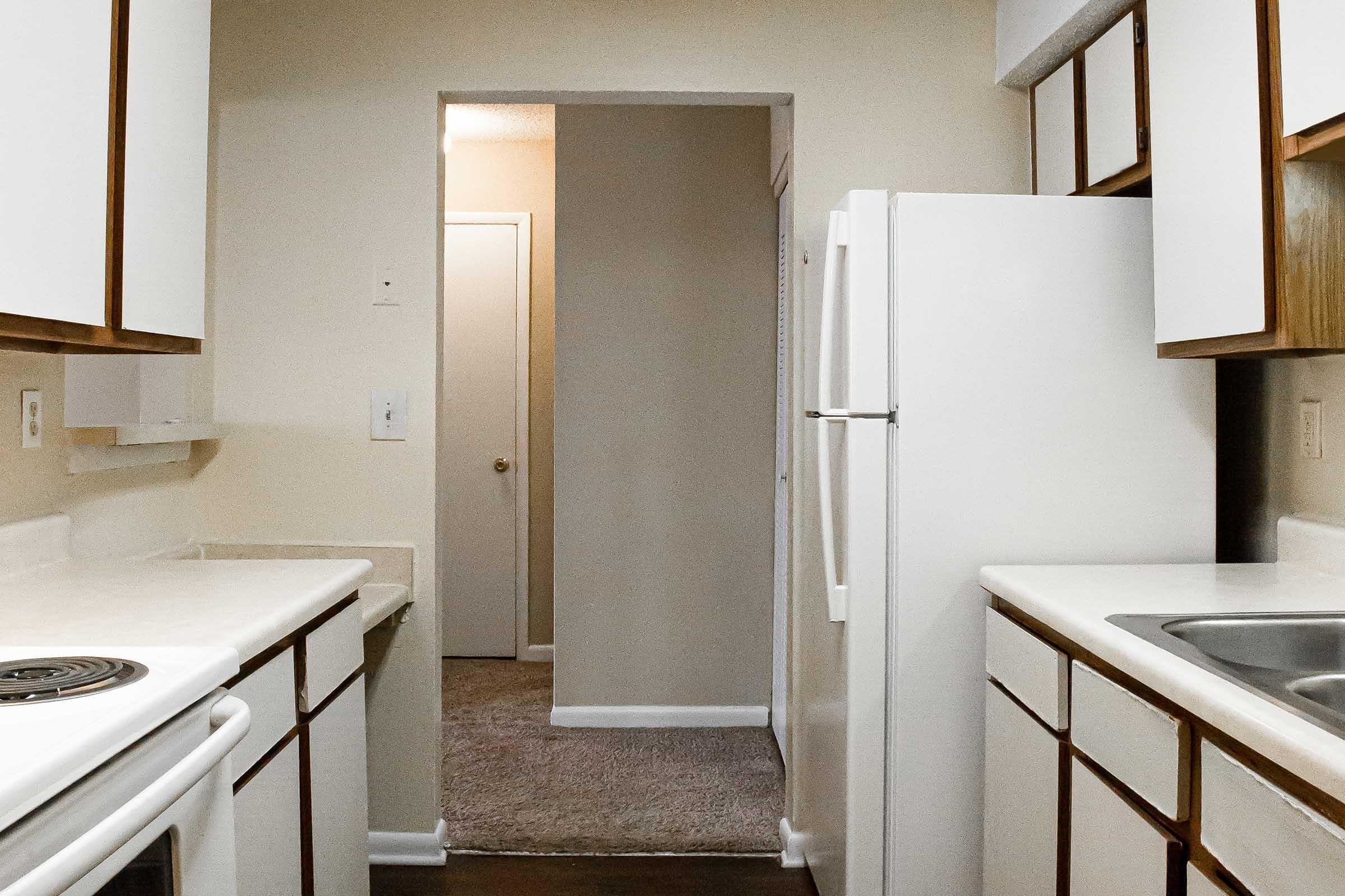 A small kitchen featuring white cabinets and countertops, a white refrigerator, a stove, and a sink. The entrance to another room is visible at the end of the kitchen, with beige walls and carpeted flooring. The space is well-lit and organized.