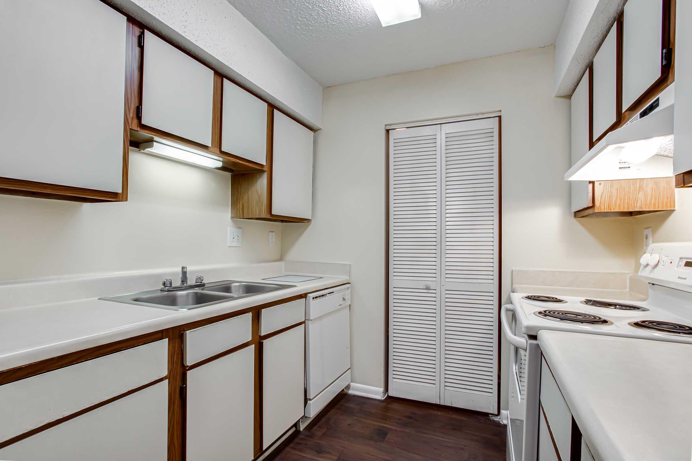 A compact kitchen featuring wooden cabinetry, a double sink, a dishwasher, and a stove with an overhead hood. The walls are light-colored, and the floor is dark, adding contrast to the space. A closed closet with folding doors is visible in the background, creating a tidy and functional environment.