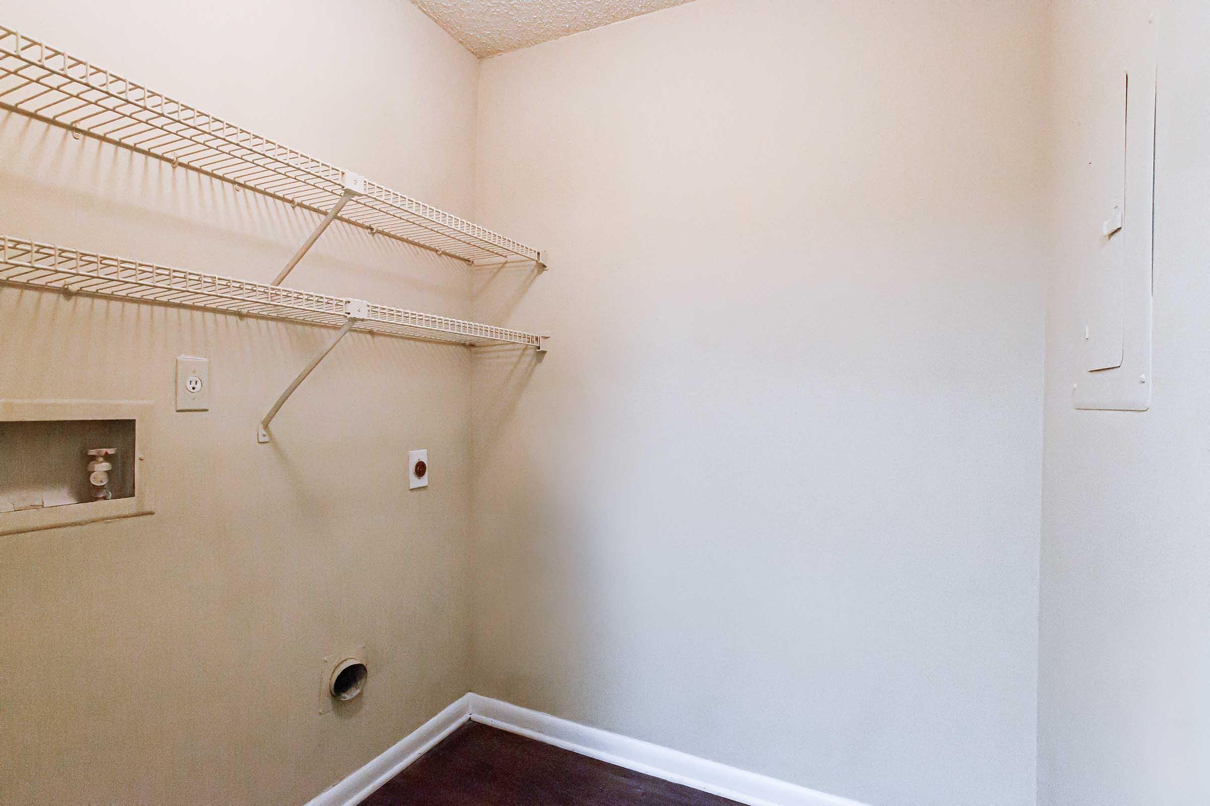 A small, empty room with light-colored walls and a wooden floor. There are two wire shelving units mounted on one wall, and a small recessed area with electrical outlets. A round vent pipe is visible near the floor, and the room has a minimalistic, uncluttered appearance.