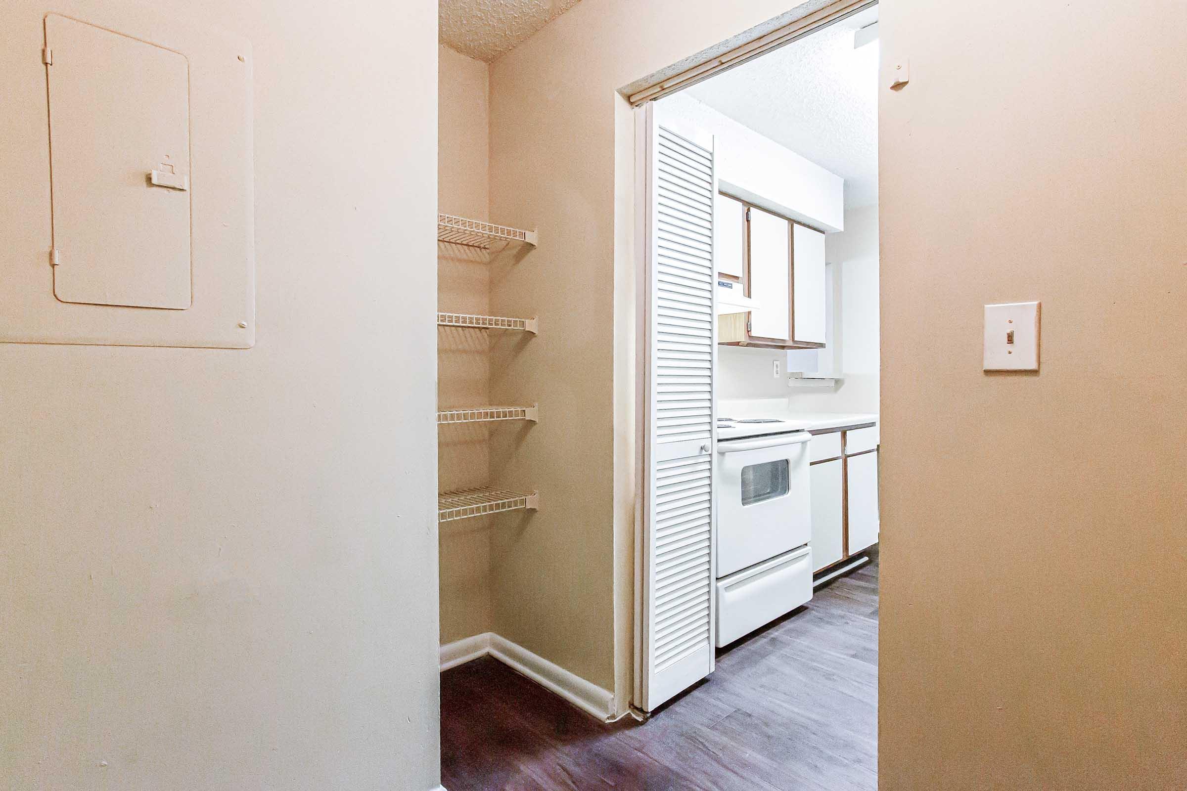 A view of a small kitchen from a hallway, featuring a white stove and cabinetry. The space has light walls, dark wood flooring, and a small shelf to the left. The entrance has a door, and there’s a utility panel on the wall. Bright lighting illuminates the room.