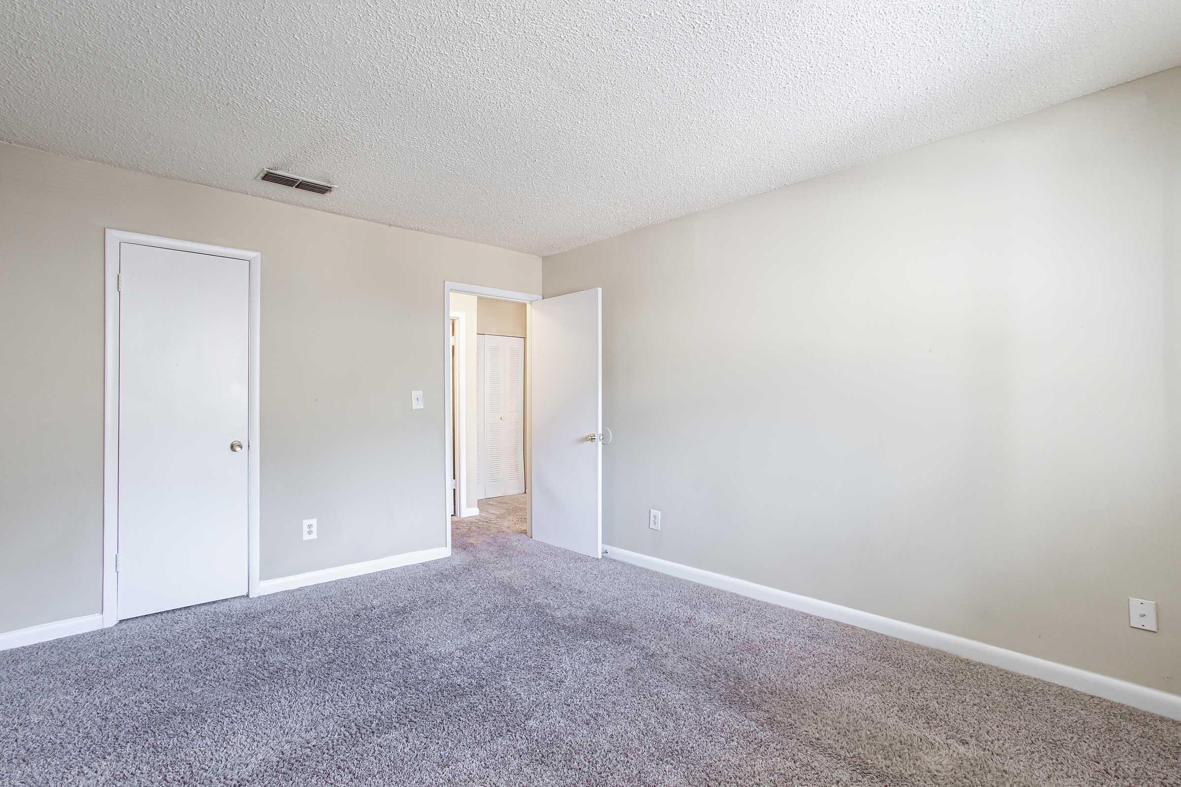 Empty room with light gray carpet, neutral-colored walls, and a closed door on the left. A second door is partially visible on the right, leading to another room. The room is well-lit with natural light flooding in, creating a spacious, inviting atmosphere.