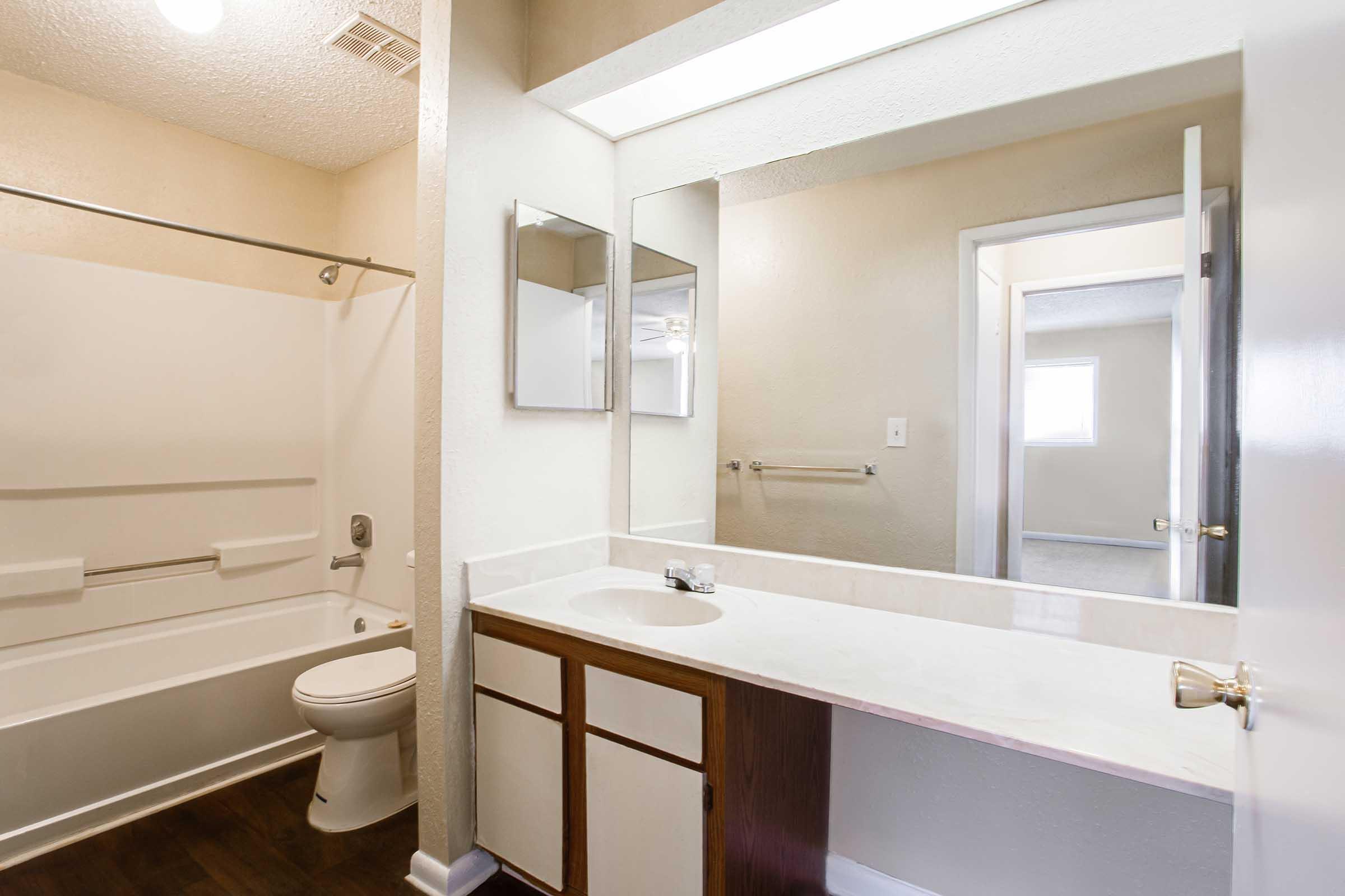 A clean, well-lit bathroom featuring a bathtub with an integrated shower, a toilet, and a double sink vanity with a mirror above. The walls are painted in a light color, and there is a door leading to another room. The overall design is modern and uncluttered.