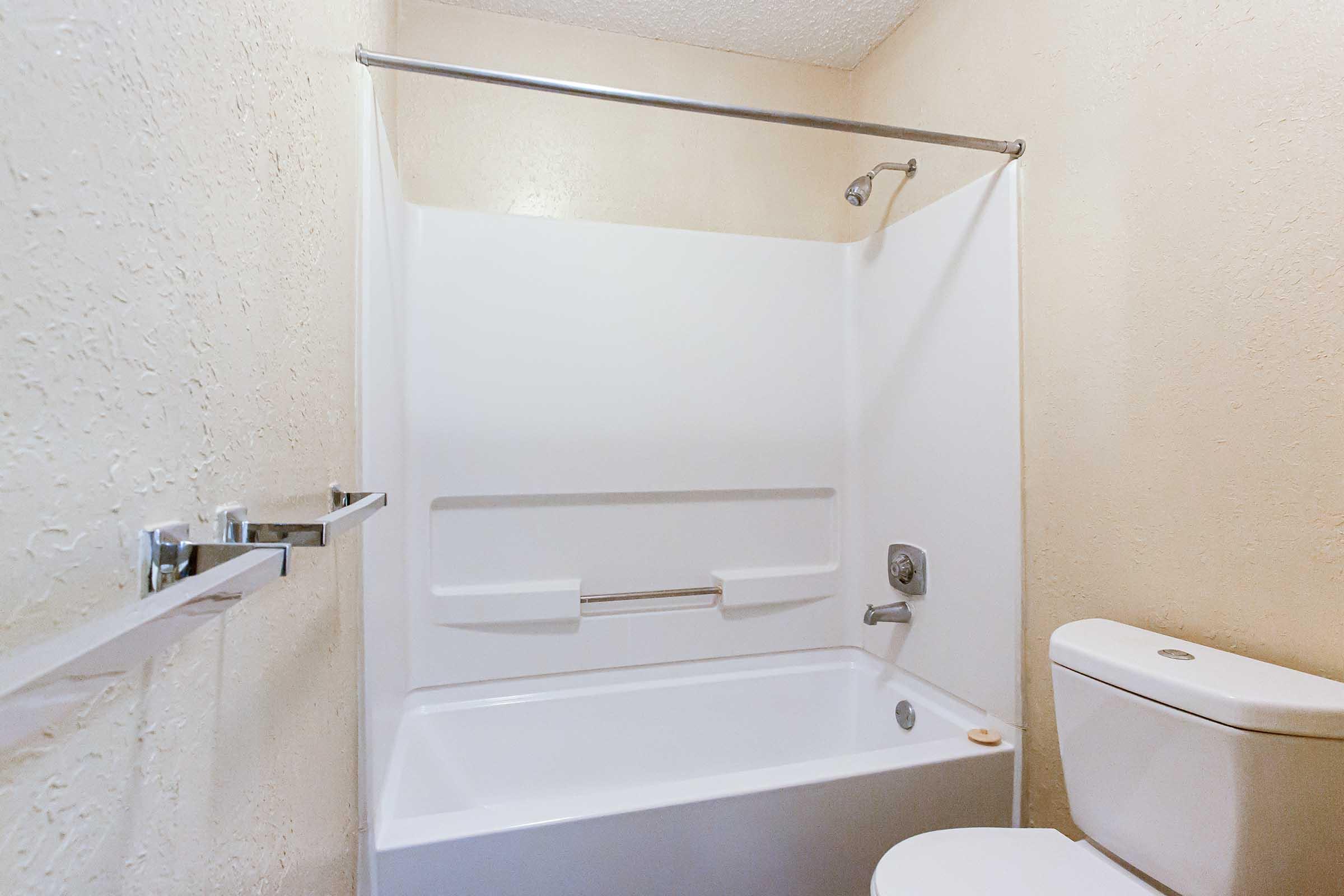 A clean, minimalistic bathroom featuring a white bathtub with an attached shower, a wall-mounted grab bar, and a white toilet. The walls are a light color, creating a bright and airy atmosphere. The flooring is not visible, but the overall design is functional and contemporary.