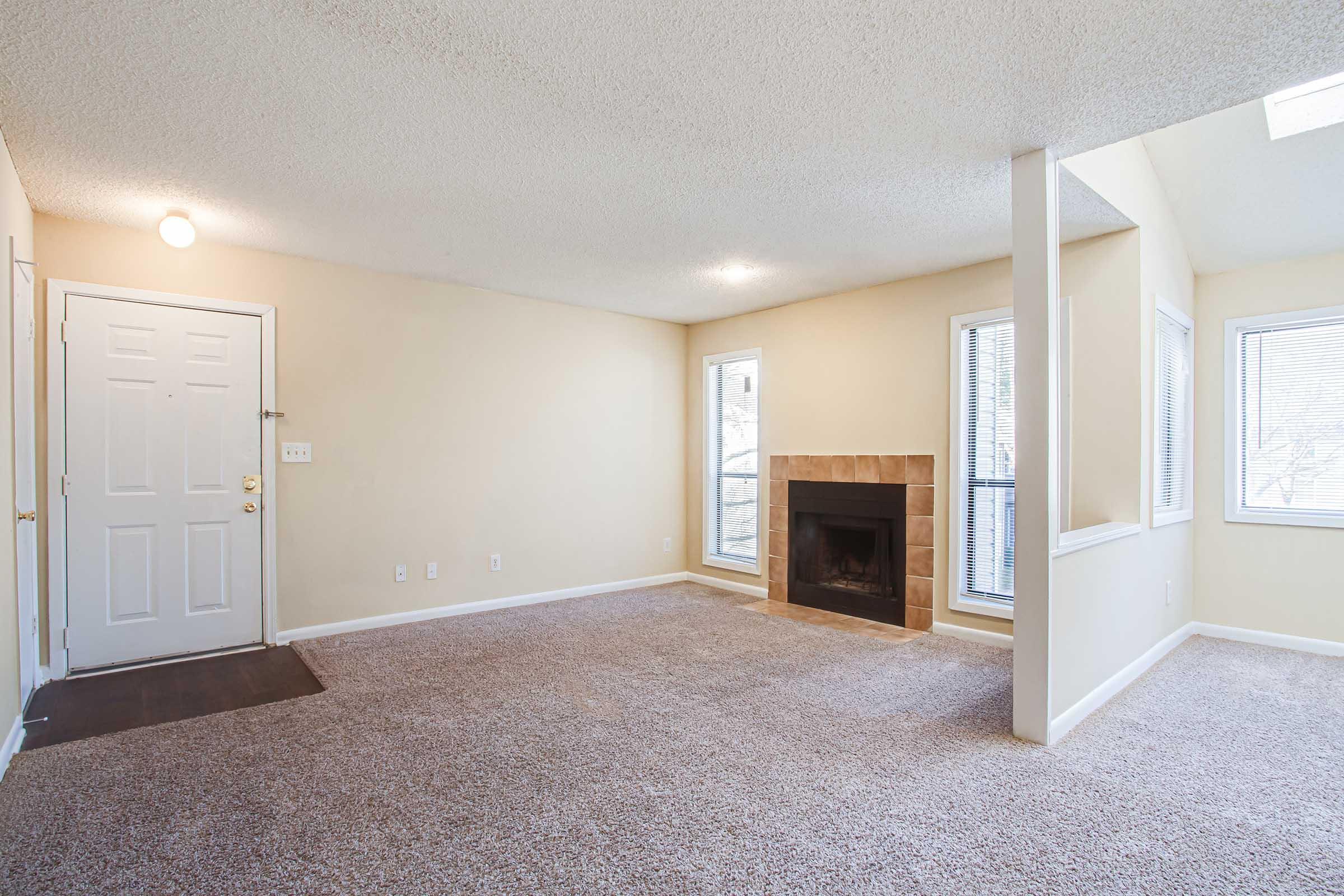 A spacious living room with light beige walls, new light fixture, and a cozy fireplace. The floor is covered with carpet, and there are windows on two sides allowing natural light. The entrance area features a door on the left and a small entryway rug. The overall ambiance is warm and inviting.