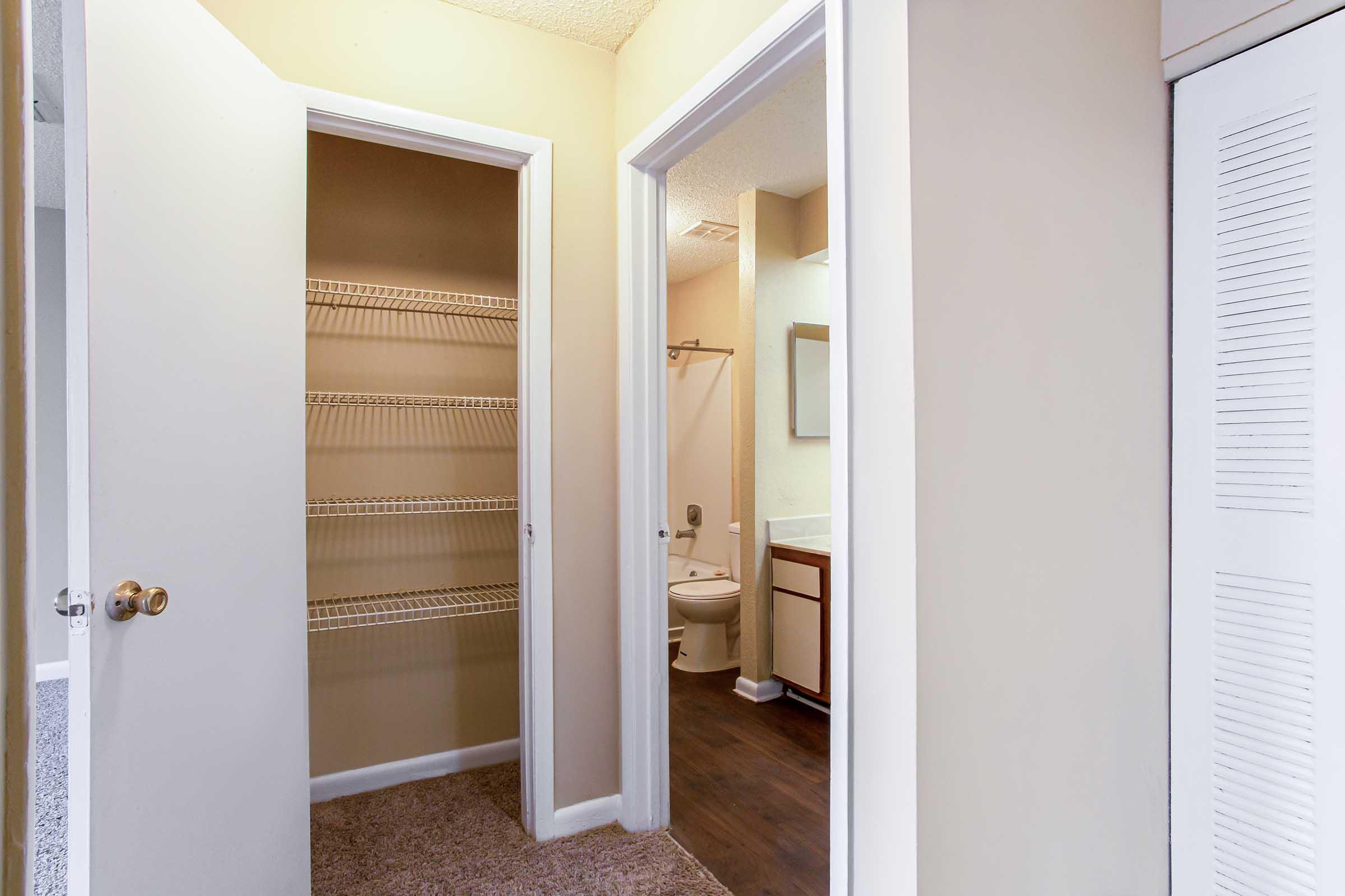 A view of a small hallway with two doorways. One doorway leads to a bathroom with a toilet and sink, while the other leads to an empty closet featuring wire shelving. The walls are painted a neutral color, and the floor is a mix of carpet and laminate. Light enters from the room ahead.