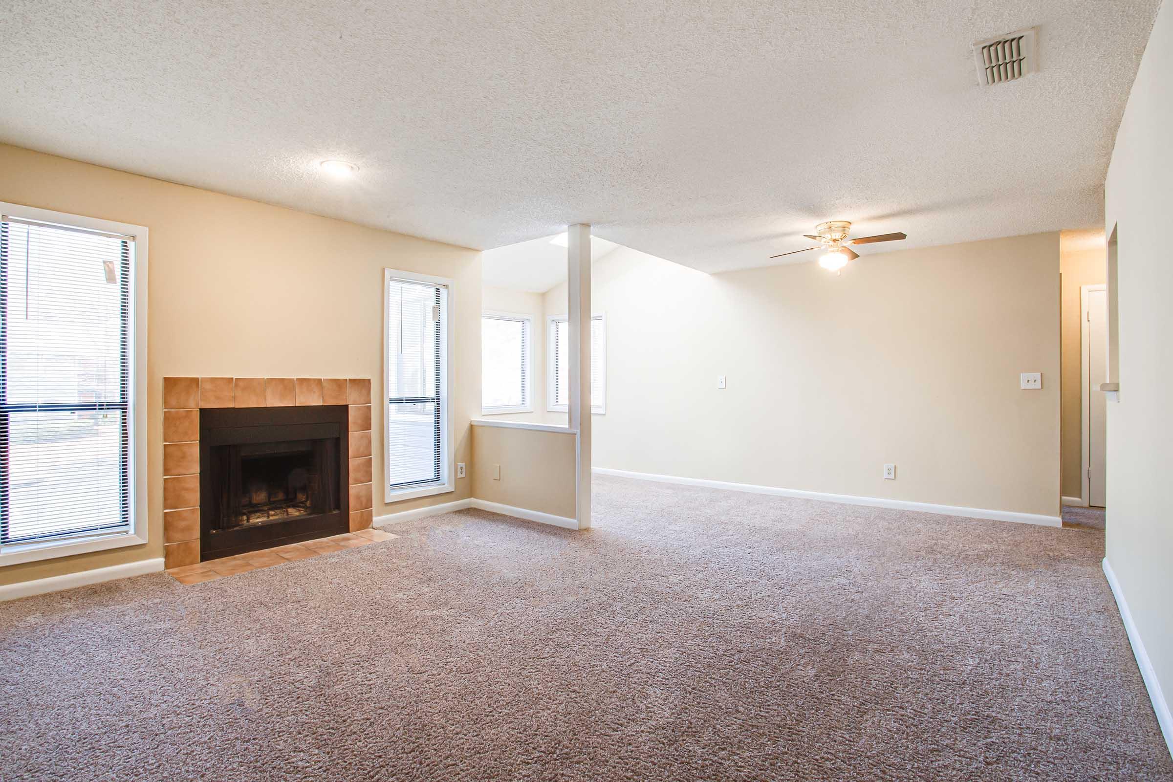A spacious living room featuring a beige color scheme, large windows allowing natural light, and a ceiling fan. There is a cozy fireplace set into a stone surround on one side, with plush carpeting covering the floor. The room appears inviting and well-maintained, with an open layout leading to other areas.