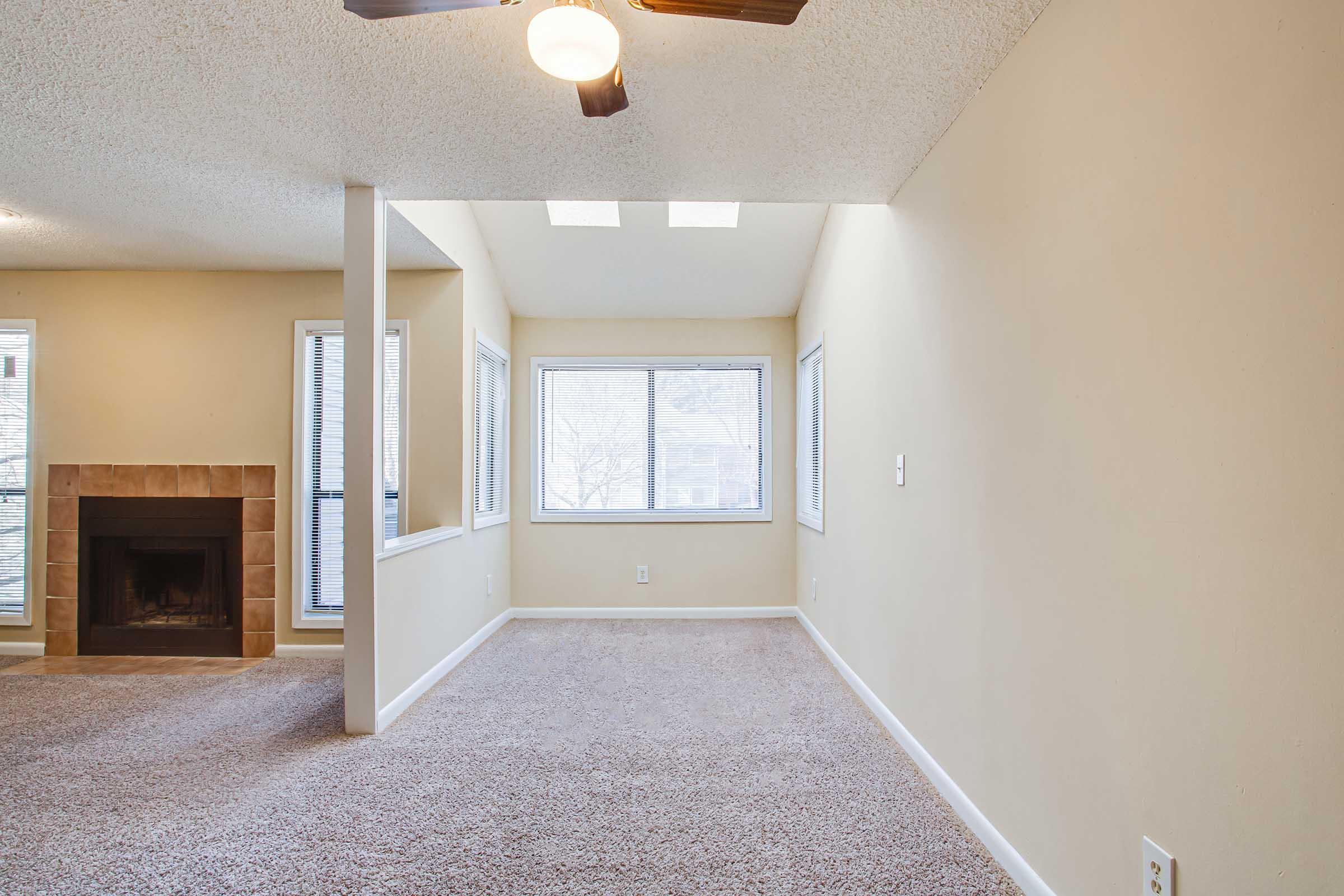 Empty living room featuring beige walls, a ceiling fan, and large windows allowing natural light. There is a beige carpeted area and a fireplace against one wall. The space is designed for comfort and brightness, with ample natural light from the windows.