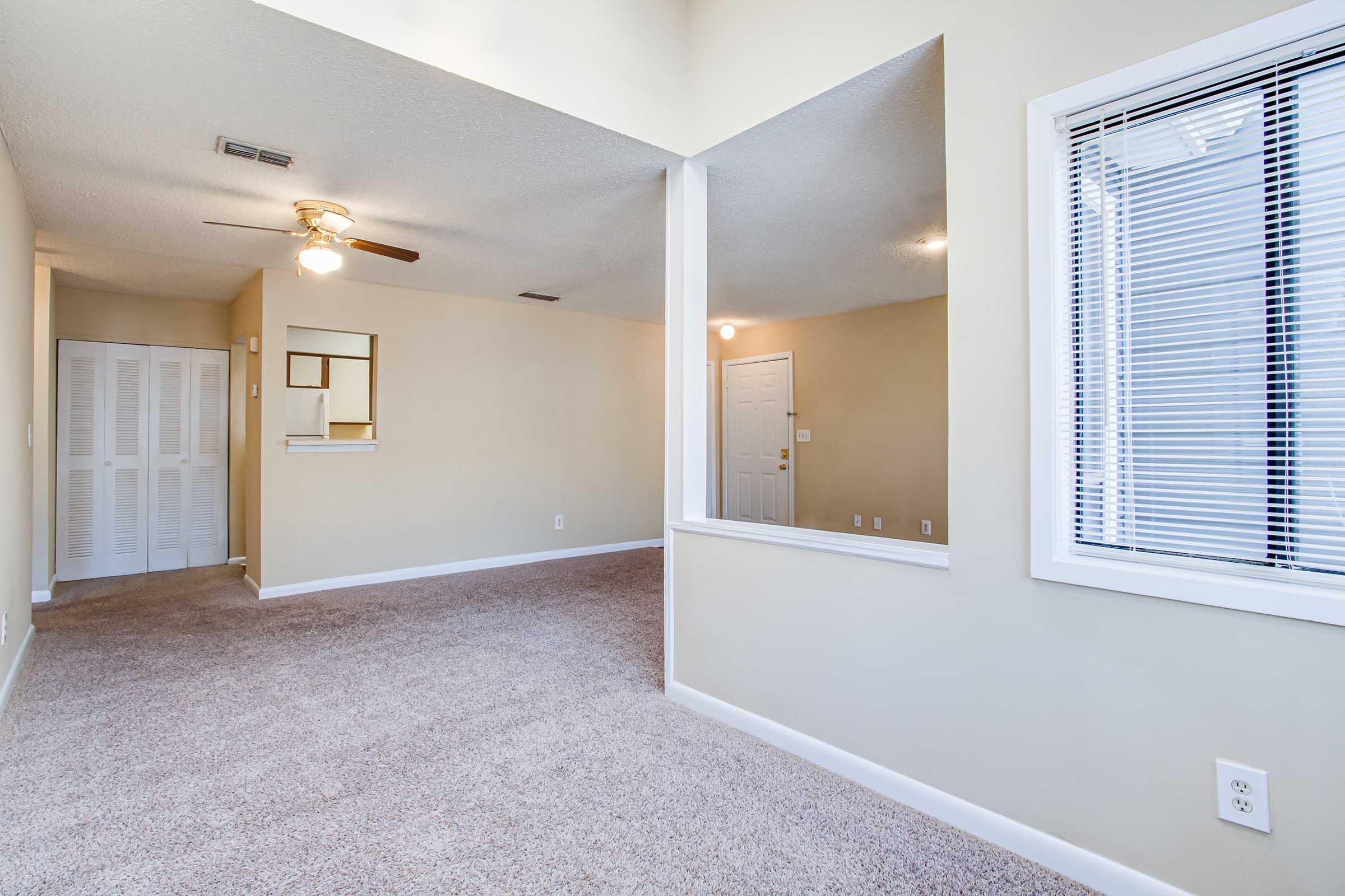 A spacious living area with beige walls and carpeted flooring. There is a ceiling fan, a small window with blinds, and a doorway leading to another room. A partial wall creates a visual separation between the living space and an adjoining area. The overall atmosphere is bright and welcoming.