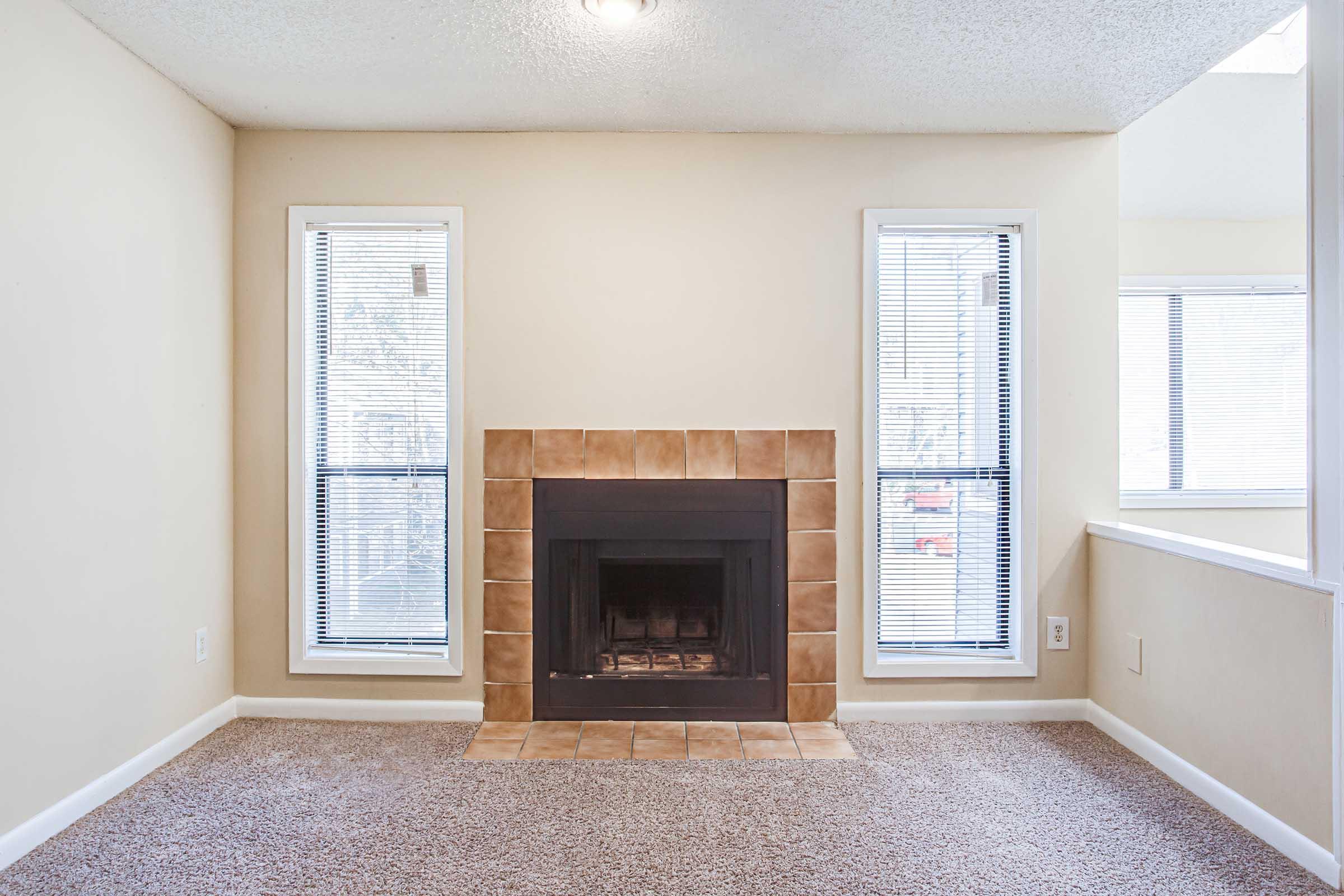 Cozy living room featuring a tiled fireplace as the focal point, flanked by two large windows with blinds. The walls are painted light beige, and the floor is covered in plush carpeting. Natural light streams in, creating a warm and inviting atmosphere.