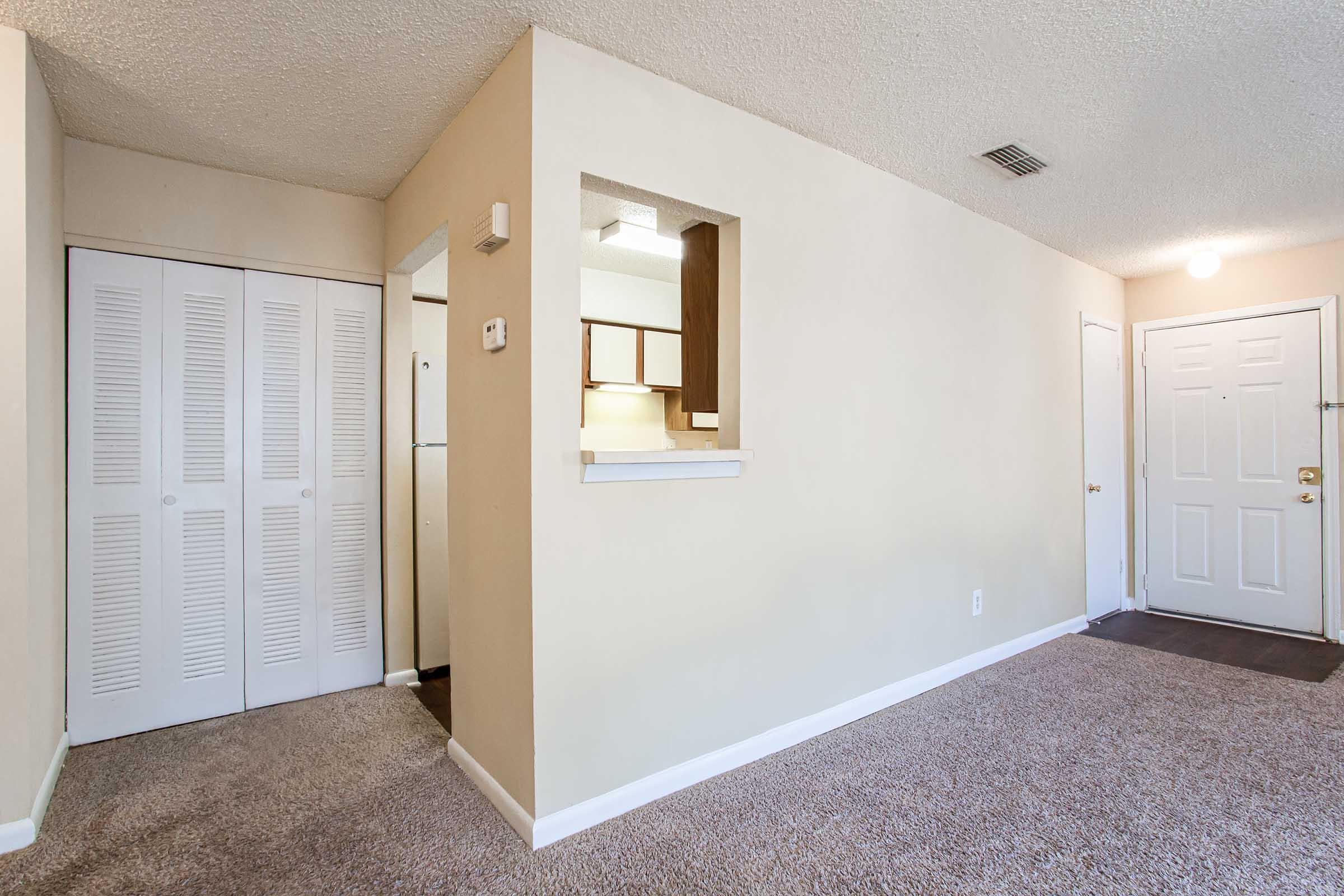 A view of an empty room featuring beige walls and carpet. On the left, there are white louvered closet doors, and to the right, a door leads outside. A small opening connects the living area to a kitchen area, which is not fully visible. Natural light enters through a ceiling light fixture.
