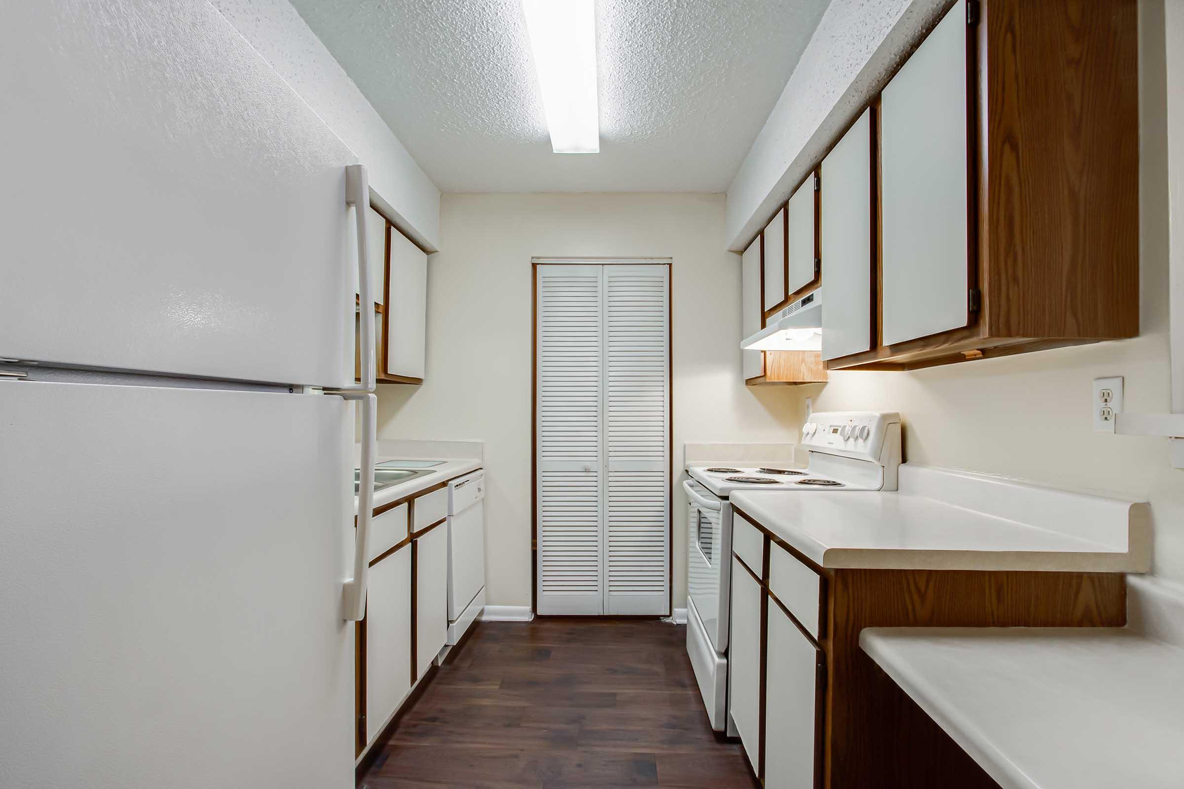 A modern kitchen featuring white appliances, light-colored cabinetry, and dark laminate flooring. The space is well-lit with a fluorescent ceiling light and includes a refrigerator, stove, and washer/dryer units. Cupboards and a closet provide ample storage. The walls are painted in a light neutral color.