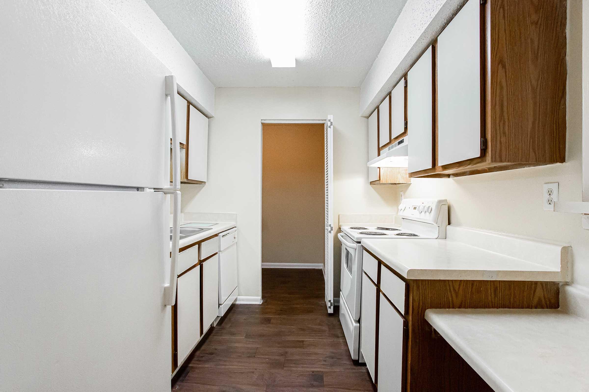 A narrow kitchen featuring white appliances, including a refrigerator, stove, and washer/dryer. Cabinets are a mix of white and wood finish, with ample counter space. The room is well-lit with overhead lighting and has a neutral-colored wall at the far end leading to a doorway.