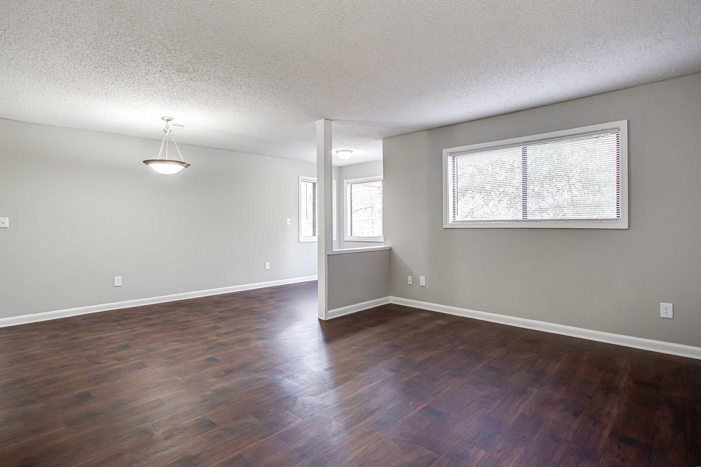 Interior view of a spacious, well-lit living room featuring a matching beige wall and dark hardwood flooring. Windows allow natural light to fill the space. A pendant light hangs from the ceiling, and there are entryways leading to adjacent rooms. The overall ambiance is modern and inviting.
