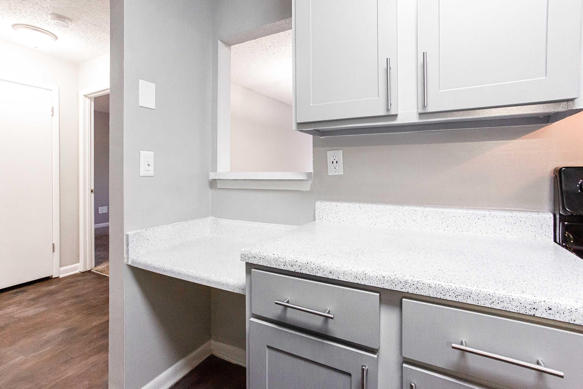A modern kitchen with gray cabinets and a white speckled countertop. There is a small cutout window leading to another room, and the walls are painted in a light gray color. The floor features dark vinyl planks, adding contrast to the bright and airy space.