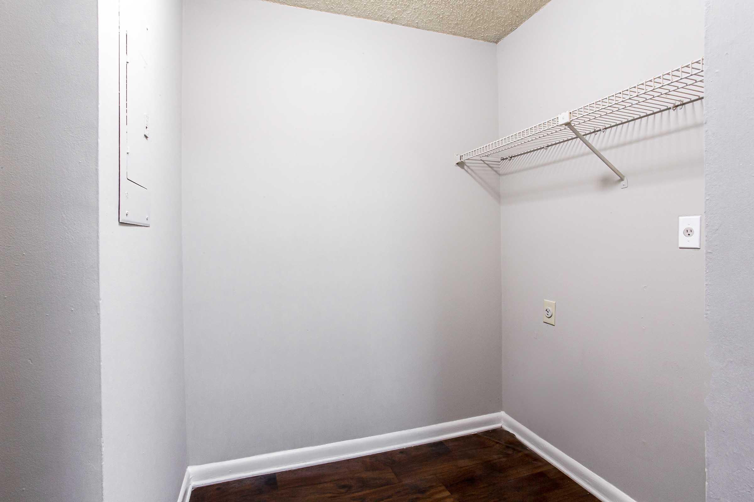 A small, empty closet featuring a light gray wall, a wire shelf mounted on one side, and a hardwood floor. The space is uncluttered and devoid of items, creating a minimalistic appearance. There is a power outlet visible on the wall.