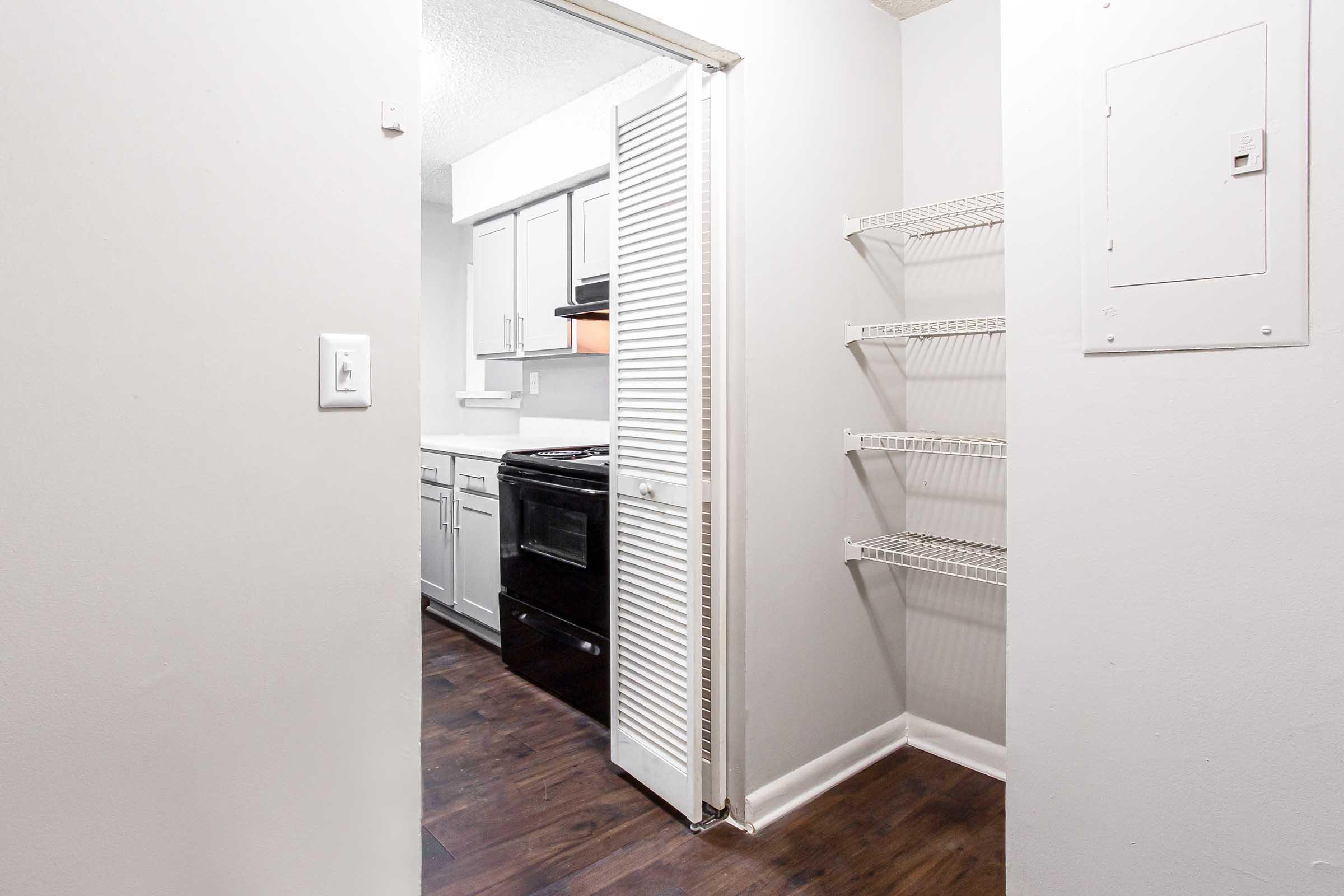 A view of a small kitchen area featuring white cabinets and a black oven. There is a doorway leading into the kitchen, and a set of wire shelves mounted on the wall. The floor is dark wood, and the overall color scheme is light and neutral.