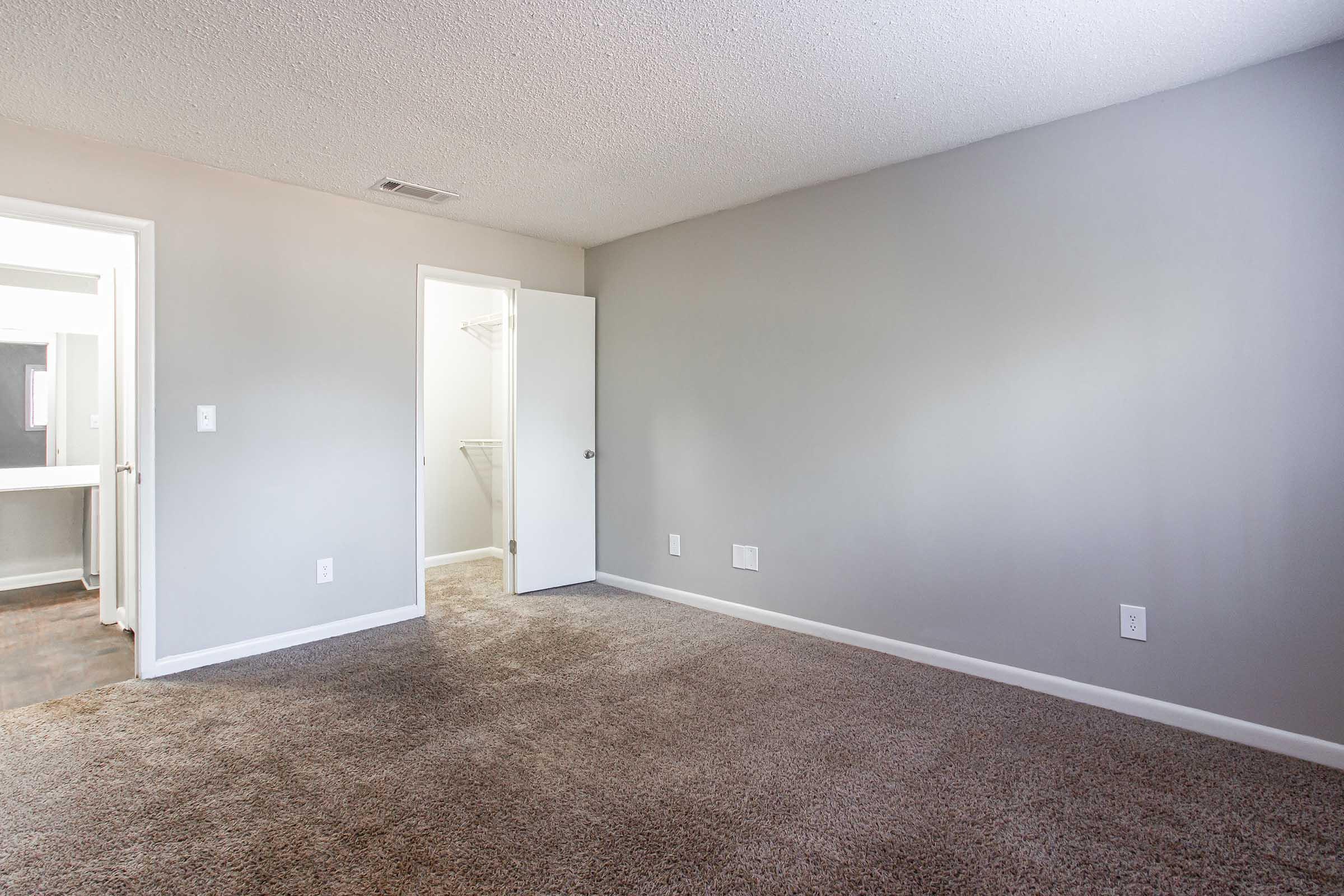 Empty room with light gray walls and brown carpet. A closed white door leads to a closet, while an open door on the left side showcases a bathroom area. The space is well-lit and uncluttered, providing a neutral backdrop suitable for various decorating styles.