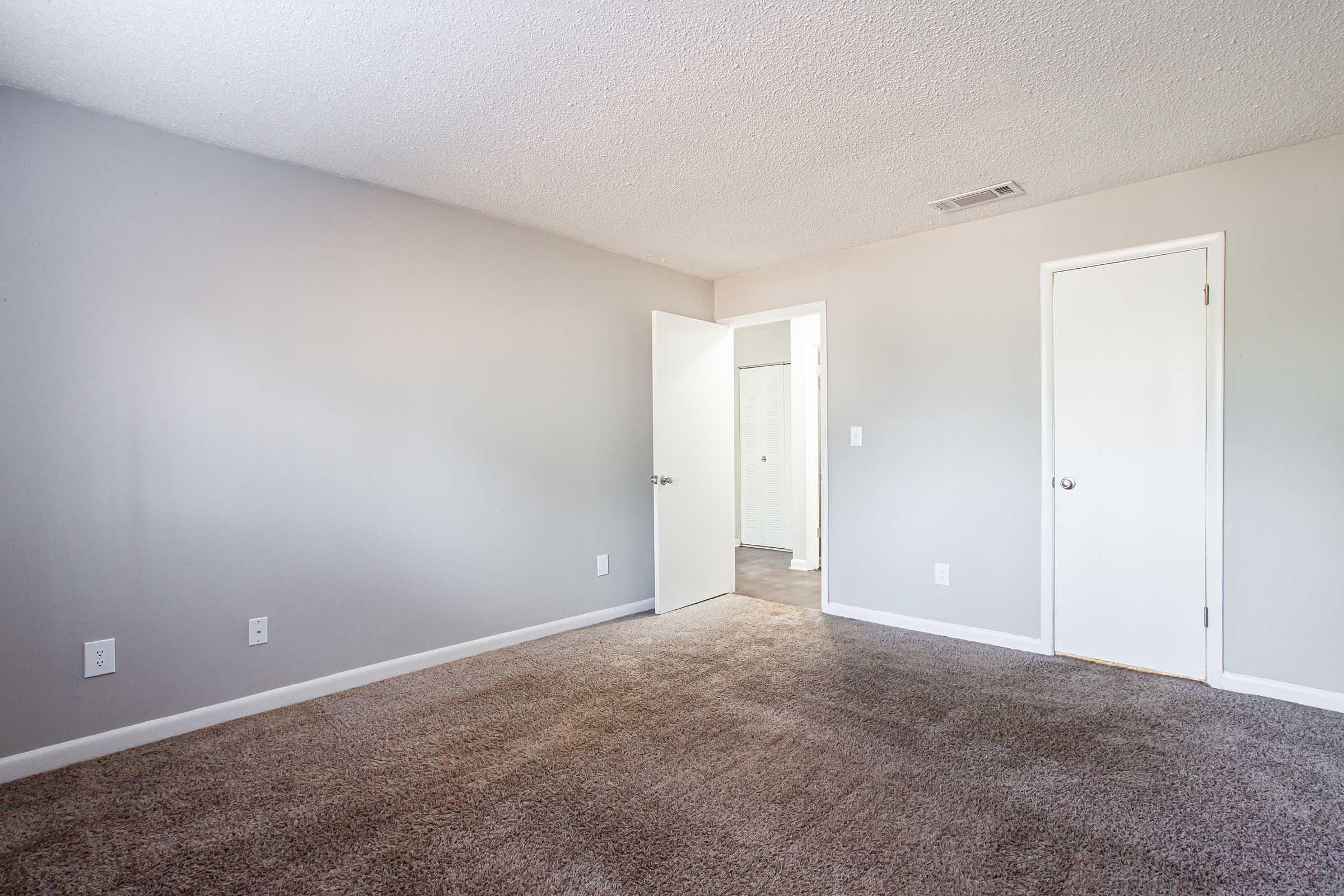 An empty room with light gray walls and carpeted flooring. A white door is open on the left, leading to another space, while a closed door is visible on the right. Natural light is coming from an unseen source, creating a bright and spacious atmosphere.