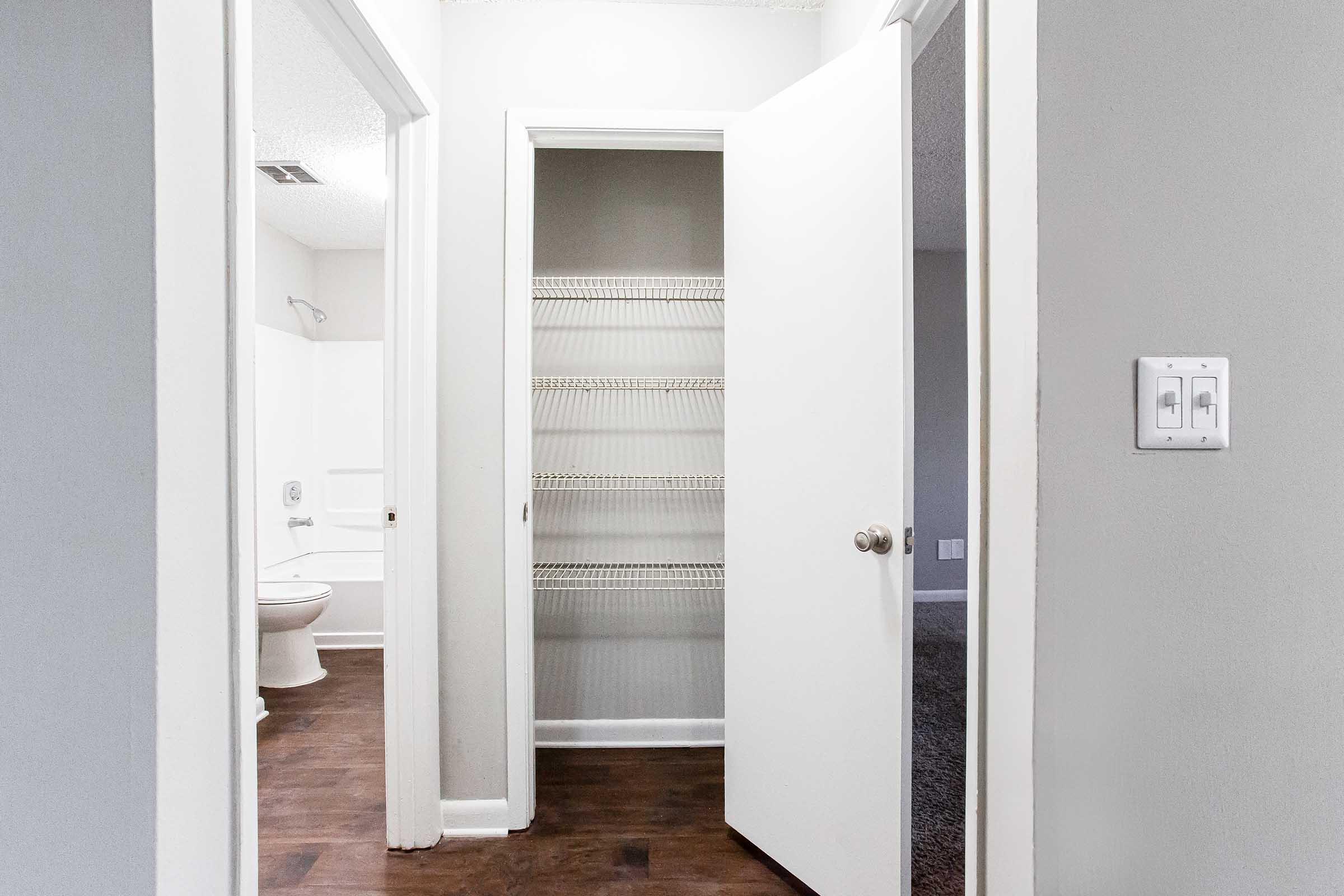 A view of a hallway with two open doors. One door leads to a closet with shelves, while the other door opens to a bathroom. The walls are painted light gray, and the flooring appears to be dark wood. The scene is well-lit, showcasing a neutral, modern aesthetic.