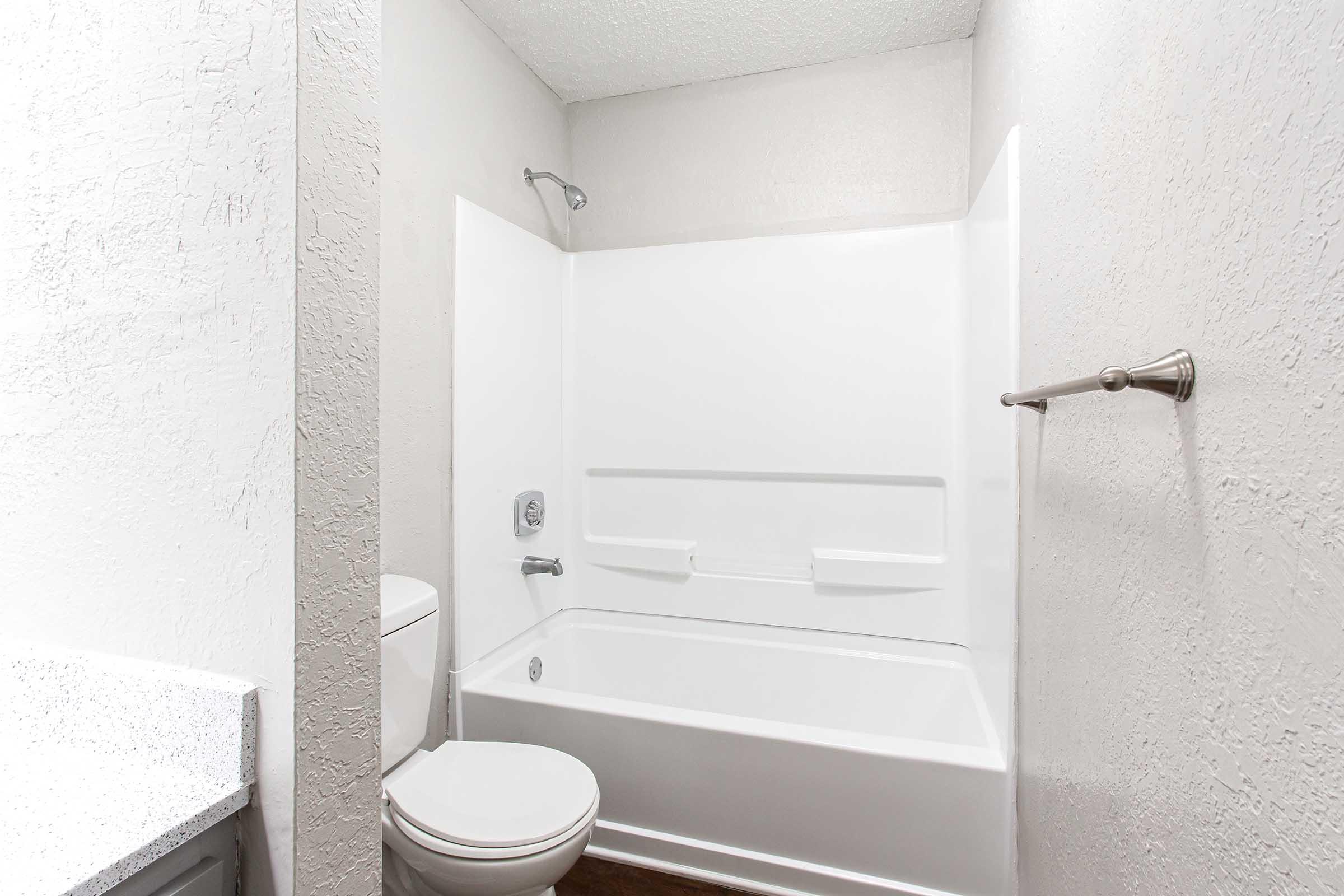 A clean, modern bathroom featuring a white bathtub with a shower, a toilet, and a light gray wall. The space includes a white countertop and a chrome towel bar, creating a bright and contemporary atmosphere.