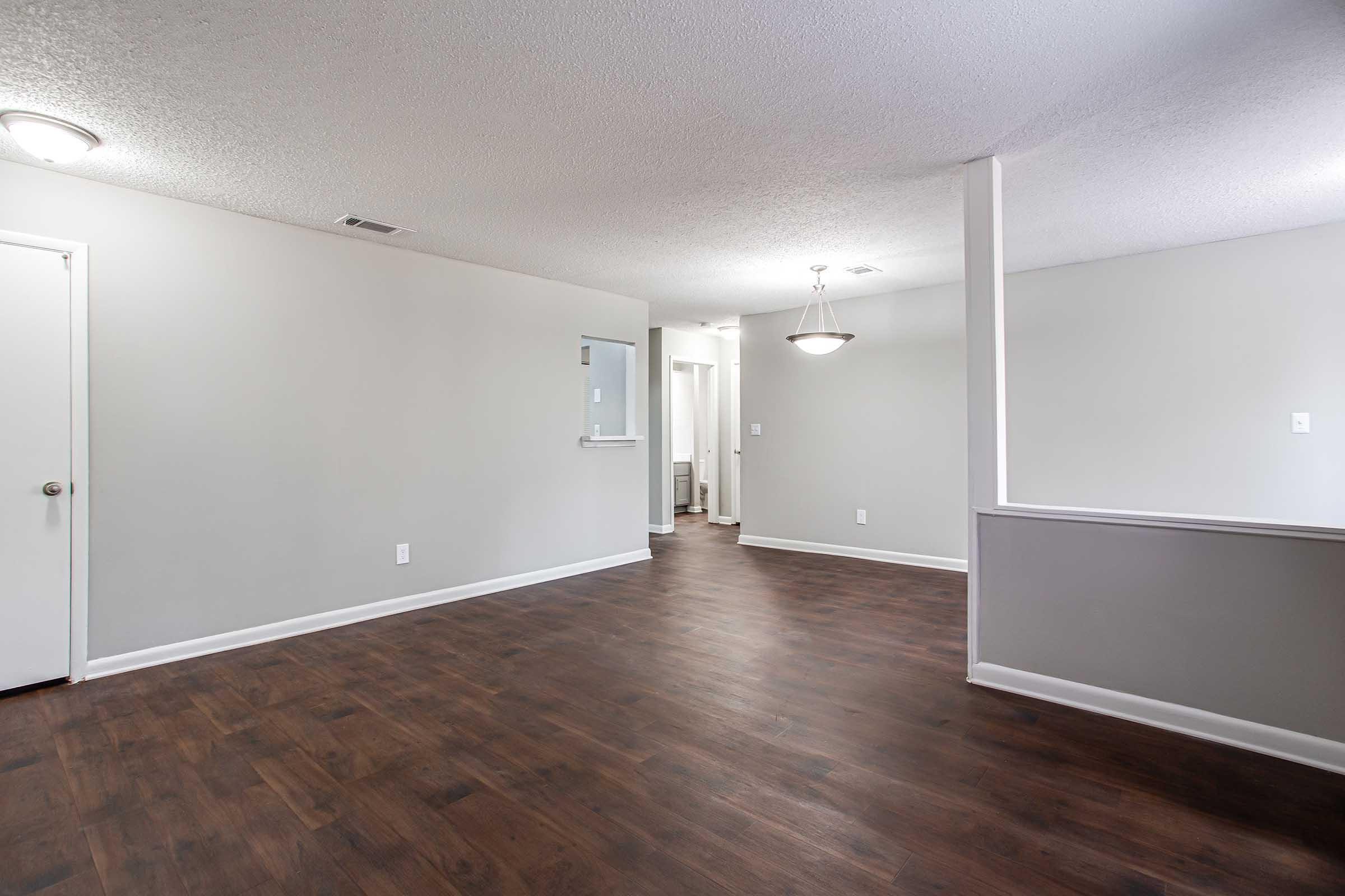 A spacious, empty interior room with light gray walls and dark wooden flooring. A light fixture hangs from the ceiling, and an open doorway leads to another area. There is a window cutout in the wall on the left, providing a view into a hallway. The overall atmosphere is bright and inviting.