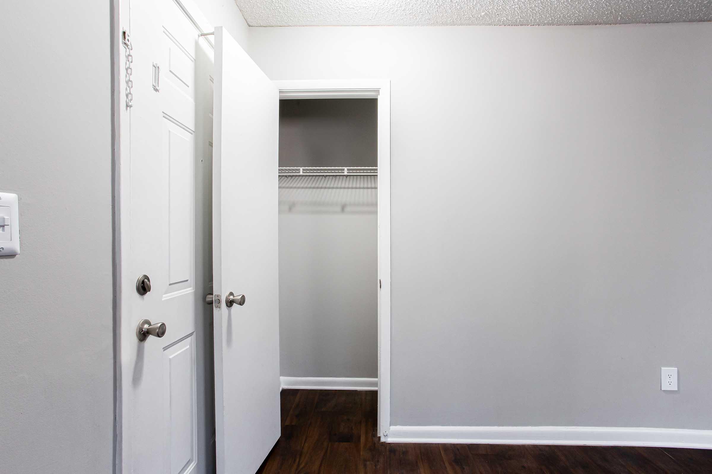 Interior view of a room featuring a white door slightly ajar, revealing a closet with a wire shelving unit. The walls are painted in a light gray color, and the floor is a dark wood finish. Natural light from a nearby window illuminates the space, giving it a clean and open appearance.