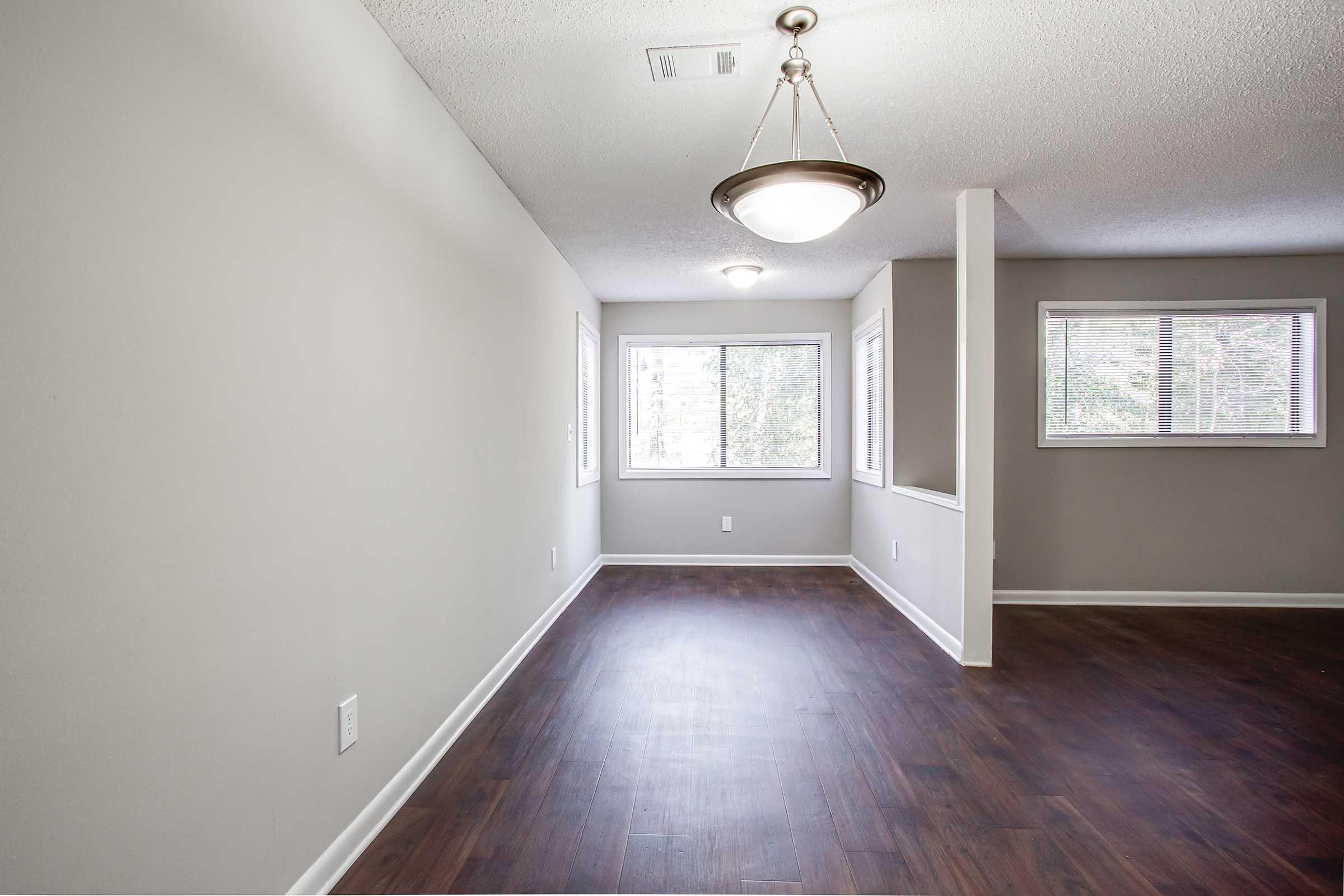 A spacious, empty room featuring light gray walls and wood laminate flooring, illuminated by a ceiling light fixture. Large windows let in natural light, and there's a small hallway visible in the background. The overall ambiance is bright and airy, suitable for various interior designs.