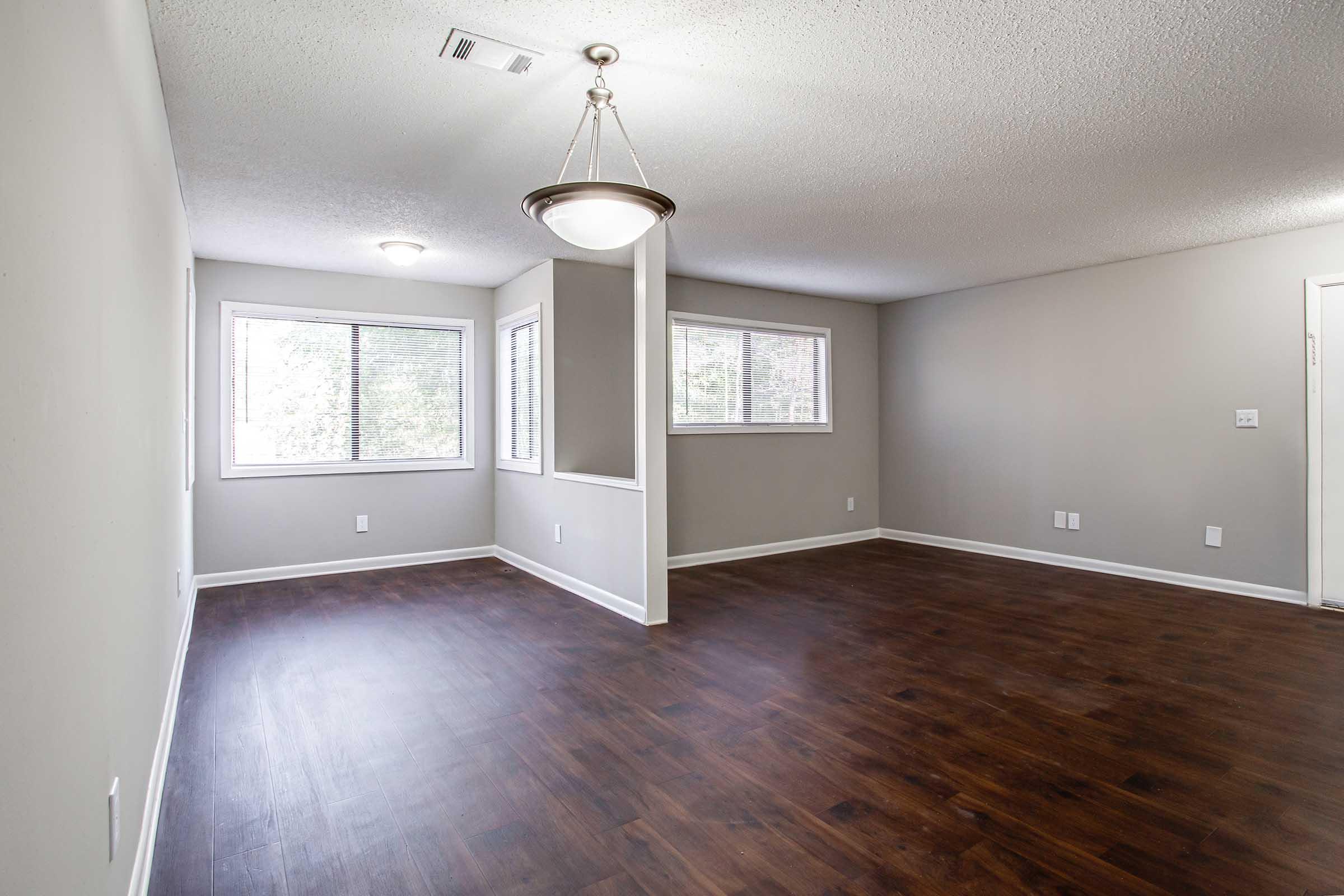 Empty interior of a modern room featuring hardwood floors, neutral-colored walls, and large windows providing natural light. A ceiling light fixture is visible, and there is a partition dividing the space. The room appears spacious and clean, suitable for various uses.