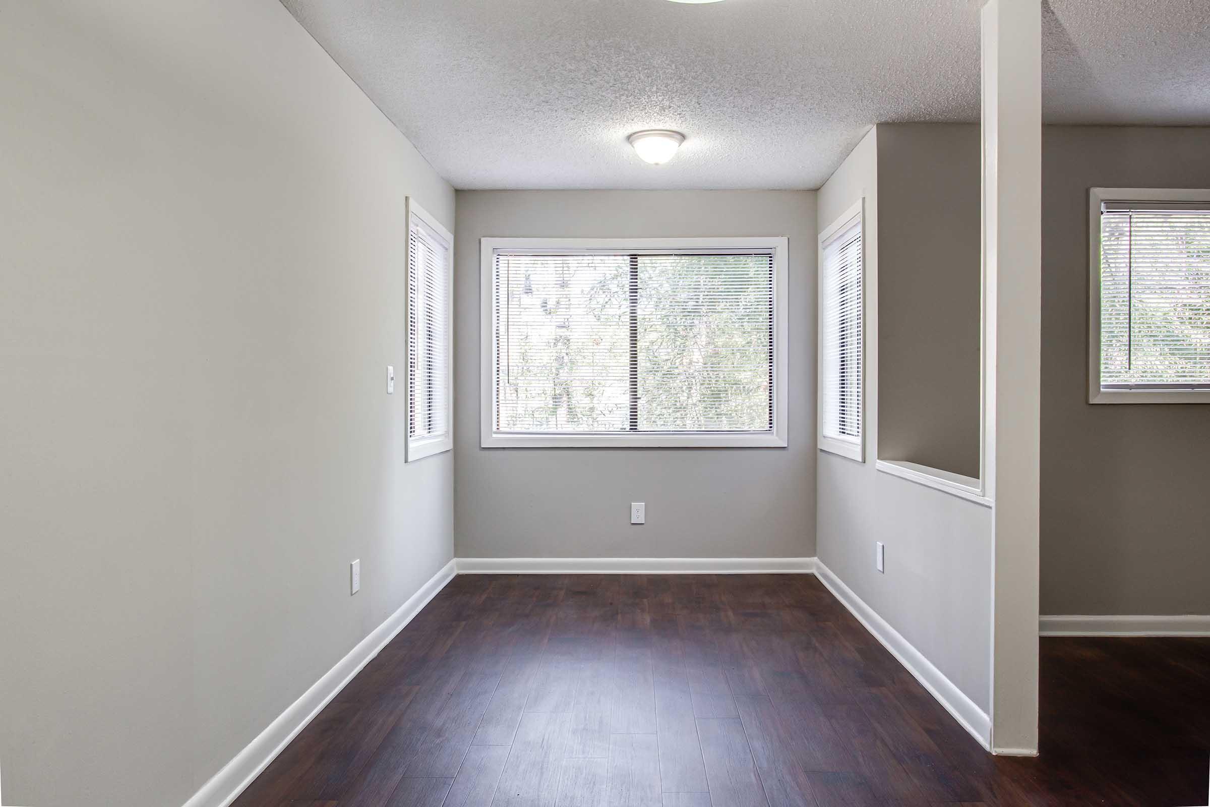 Interior of a vacant room featuring light gray walls, dark laminate flooring, and a window with blinds providing natural light. A ceiling light fixture is visible, and there is a small partition dividing the space. The room appears clean and ready for occupancy.