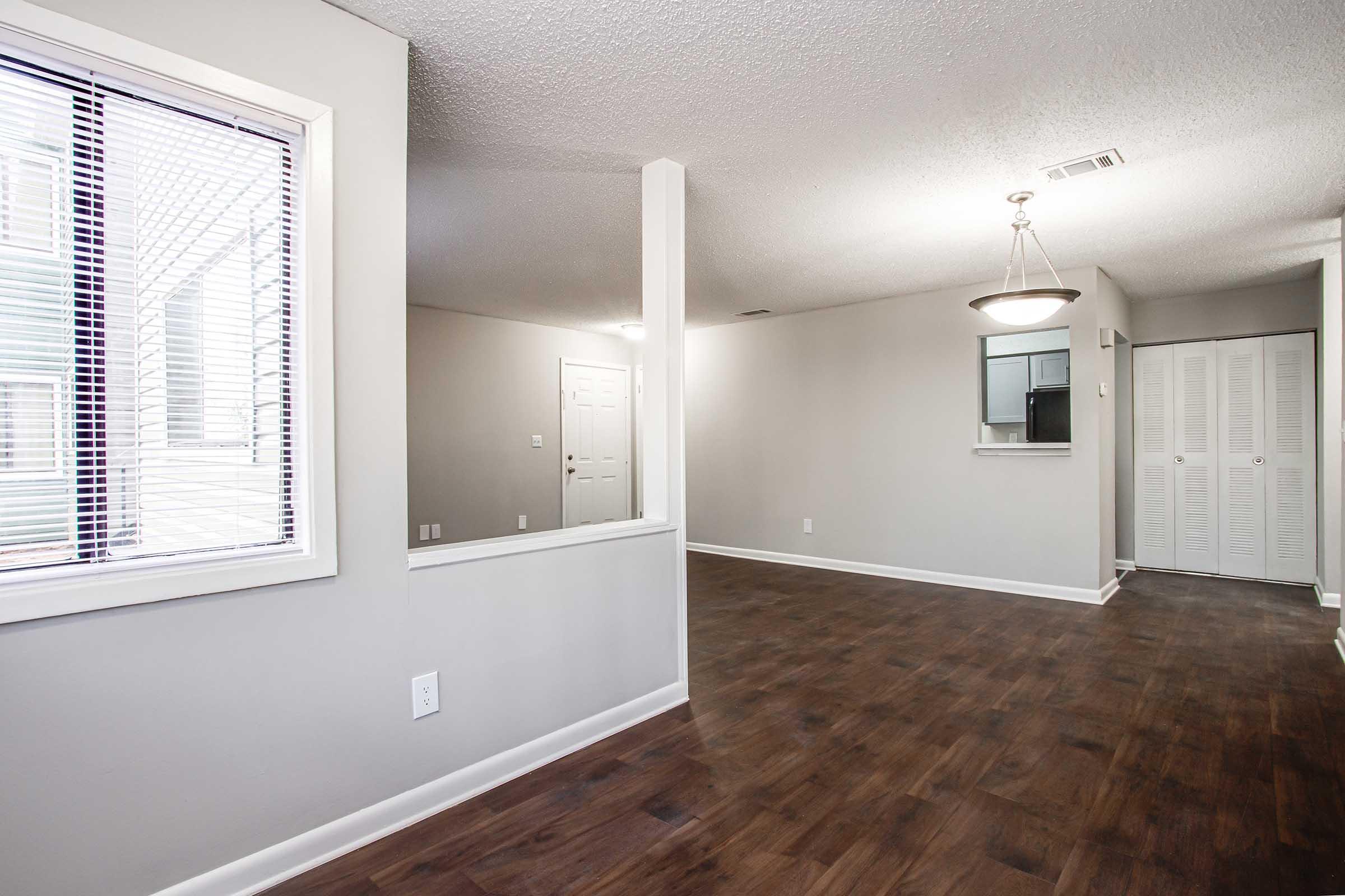 Interior view of a spacious, modern apartment. The room features light-colored walls, dark wooden flooring, and a large window with blinds. There’s an open layout with a partition wall, a ceiling light fixture, and a doorway leading to another area. A closet with doors is visible in the background.