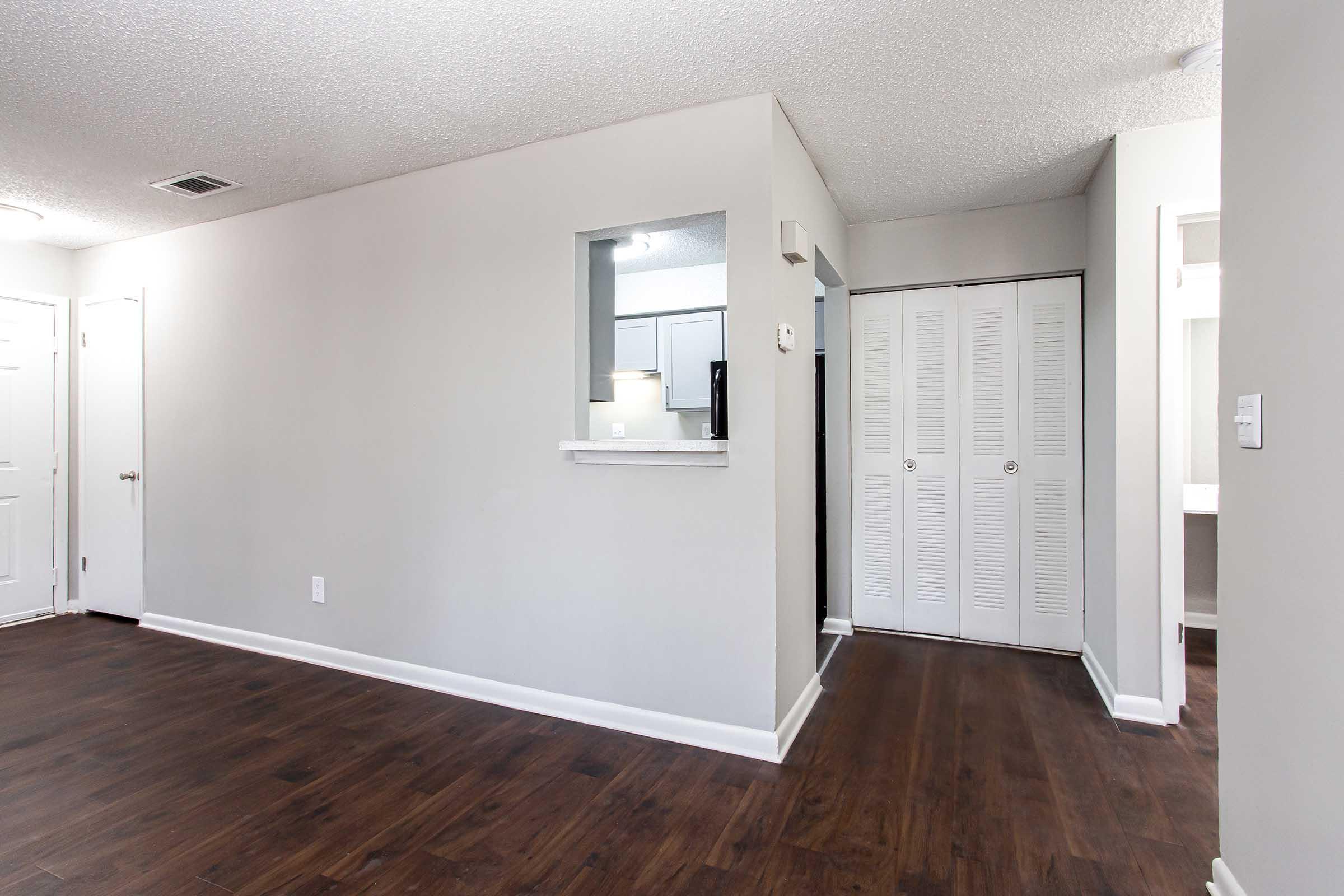 A spacious, modern interior of an apartment featuring a neutral color palette, with a textured ceiling, hardwood-like flooring, and a small service bar area. There is a doorway leading to an adjacent room and a closet with sliding doors, offering a bright and inviting atmosphere.