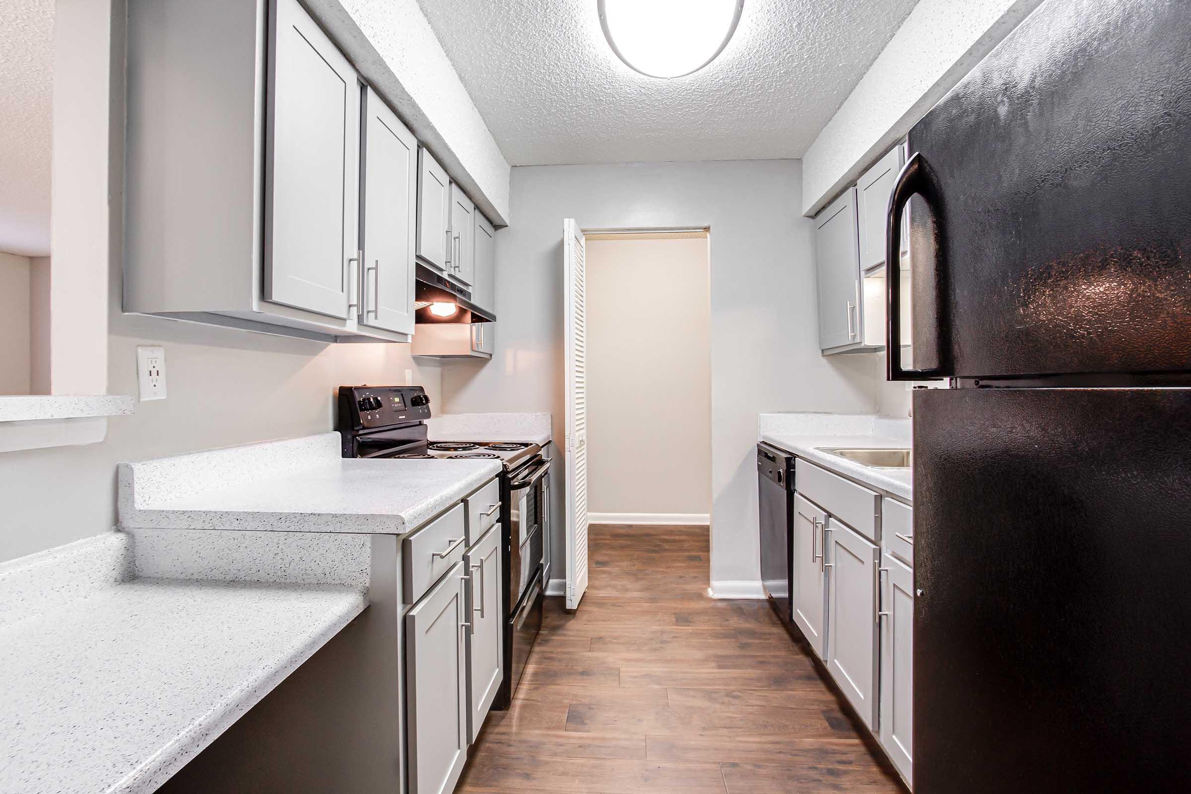 A modern kitchen featuring gray cabinets, a black refrigerator, and stainless steel appliances. The countertops are light-colored with a speckled design. There is a spacious layout with wooden flooring, providing a clean and contemporary look. The room is well-lit by a ceiling light.