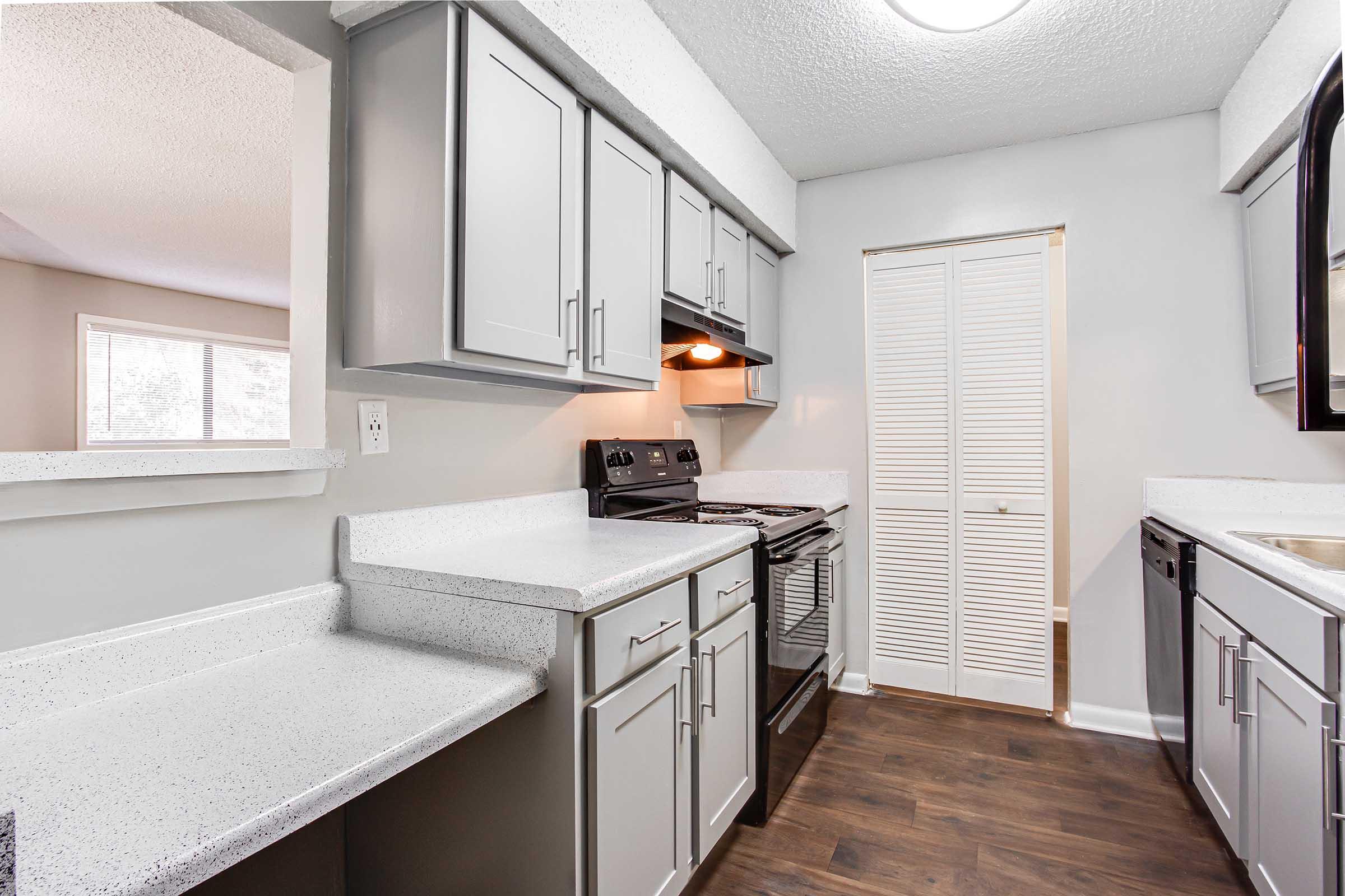 Modern kitchen interior featuring light gray cabinets, a stainless steel stove, and a black microwave. The countertop is a speckled white material, and there is a sliding door closet. Natural light comes through a window, illuminating the space. The flooring is dark wood, creating a nice contrast with the cabinetry.