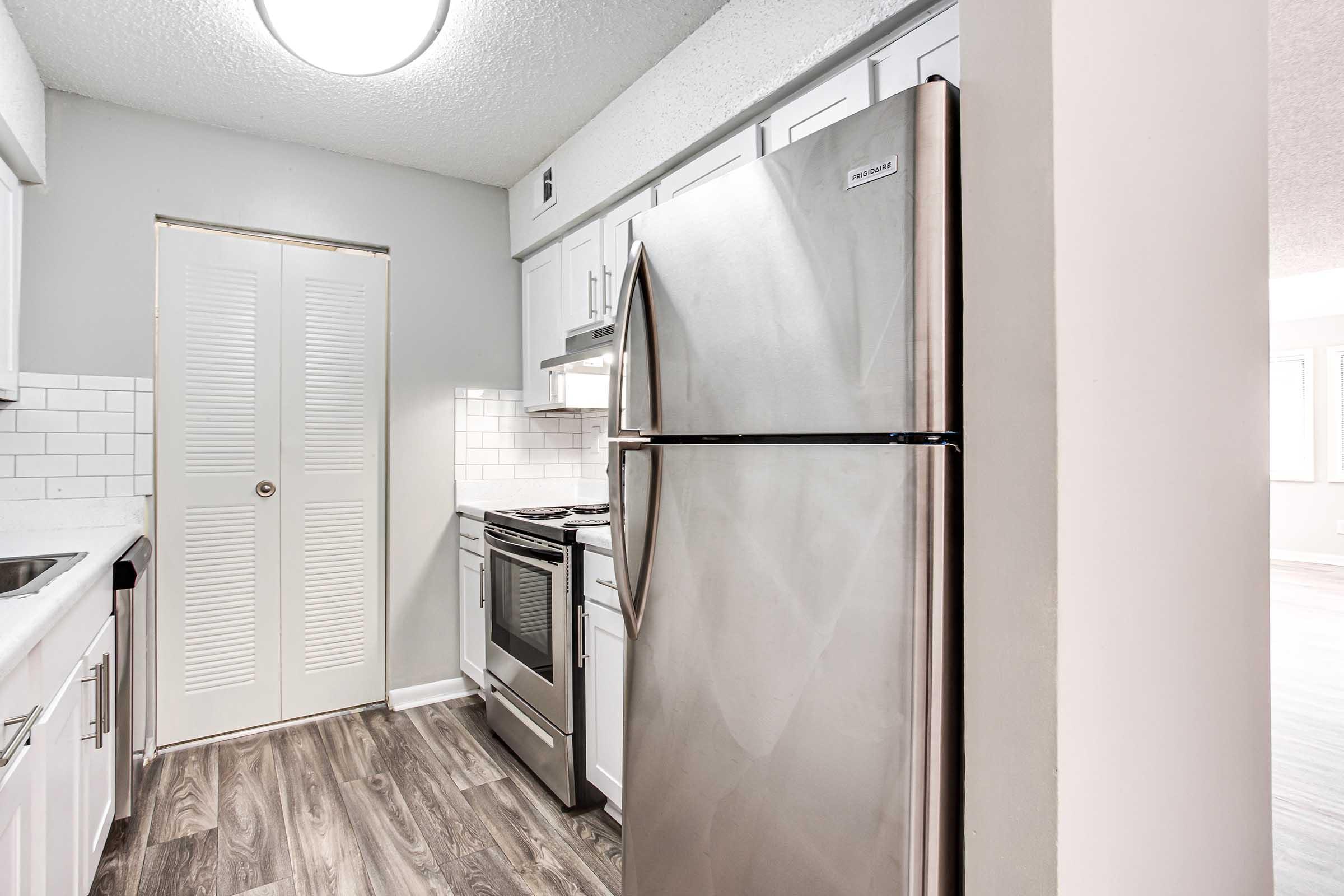 Modern kitchen featuring stainless steel appliances, including a refrigerator and oven, white cabinets, and a tiled backsplash. The space has laminate flooring and neutral-colored walls, with a door leading to a closet or pantry. Bright lighting enhances the clean and contemporary design.