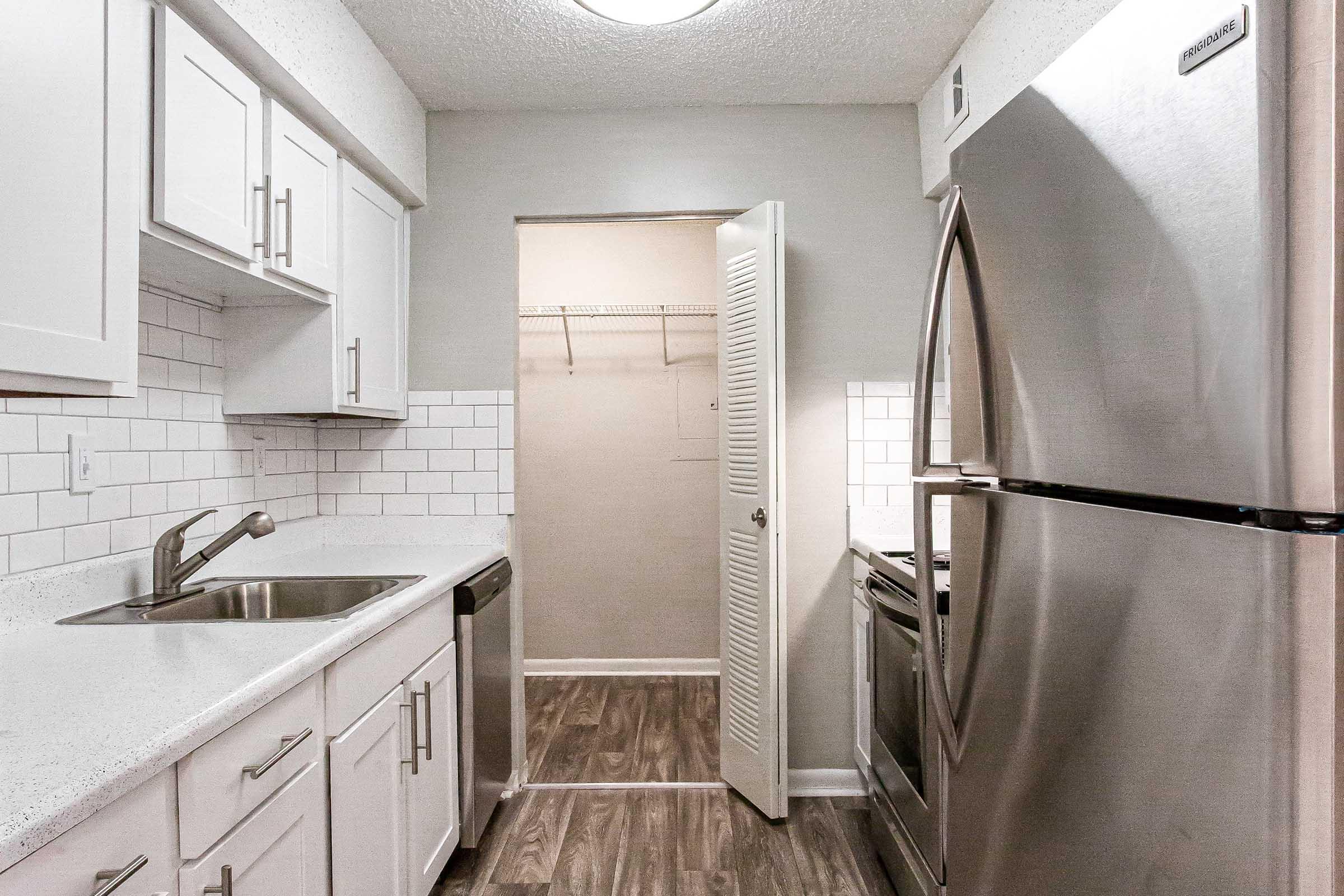 Modern kitchen featuring white cabinetry, stainless steel appliances, and a light grey color scheme. The space includes a sink with a faucet, a refrigerator, and an open closet area with shelves. The flooring is a dark wood look, enhancing the contemporary design.