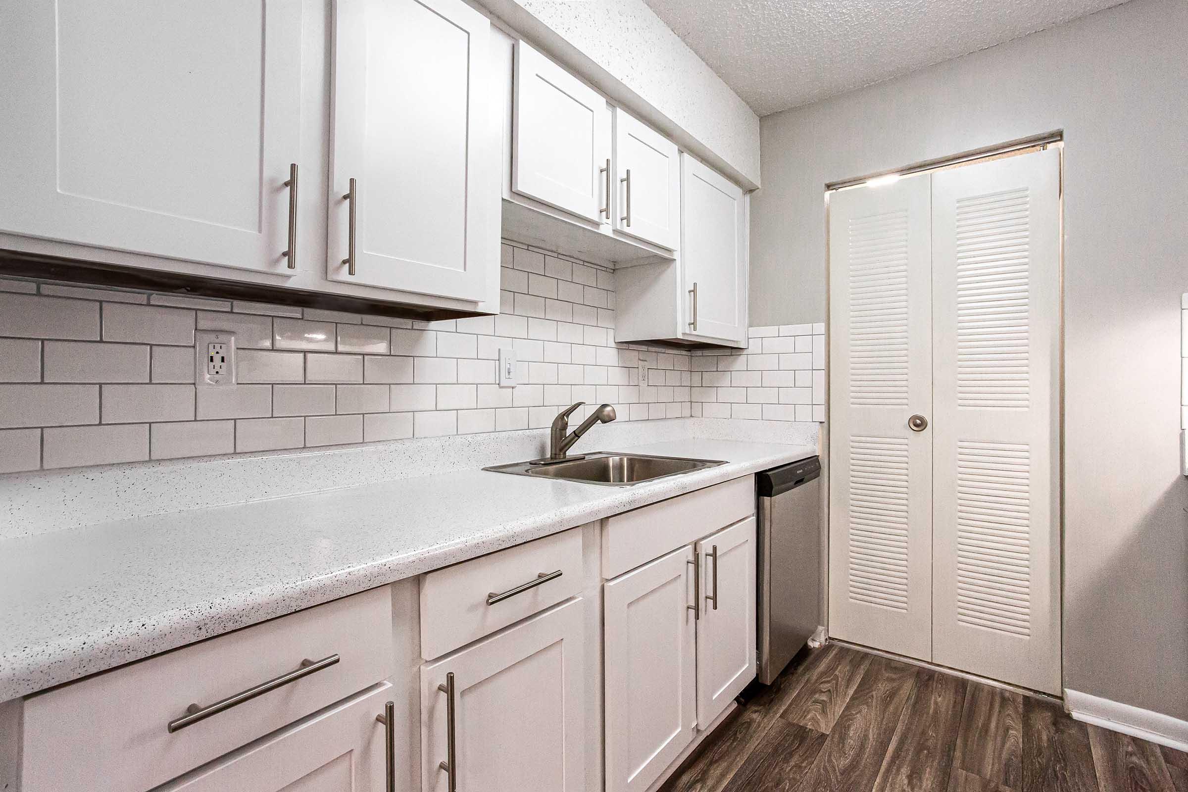 Modern kitchen with white cabinets and a light gray backsplash featuring rectangular tiles. A stainless steel sink is installed on a white countertop, and a dishwasher is visible. There's a door with louvered panels on the right side, and the flooring has a wood-like finish.