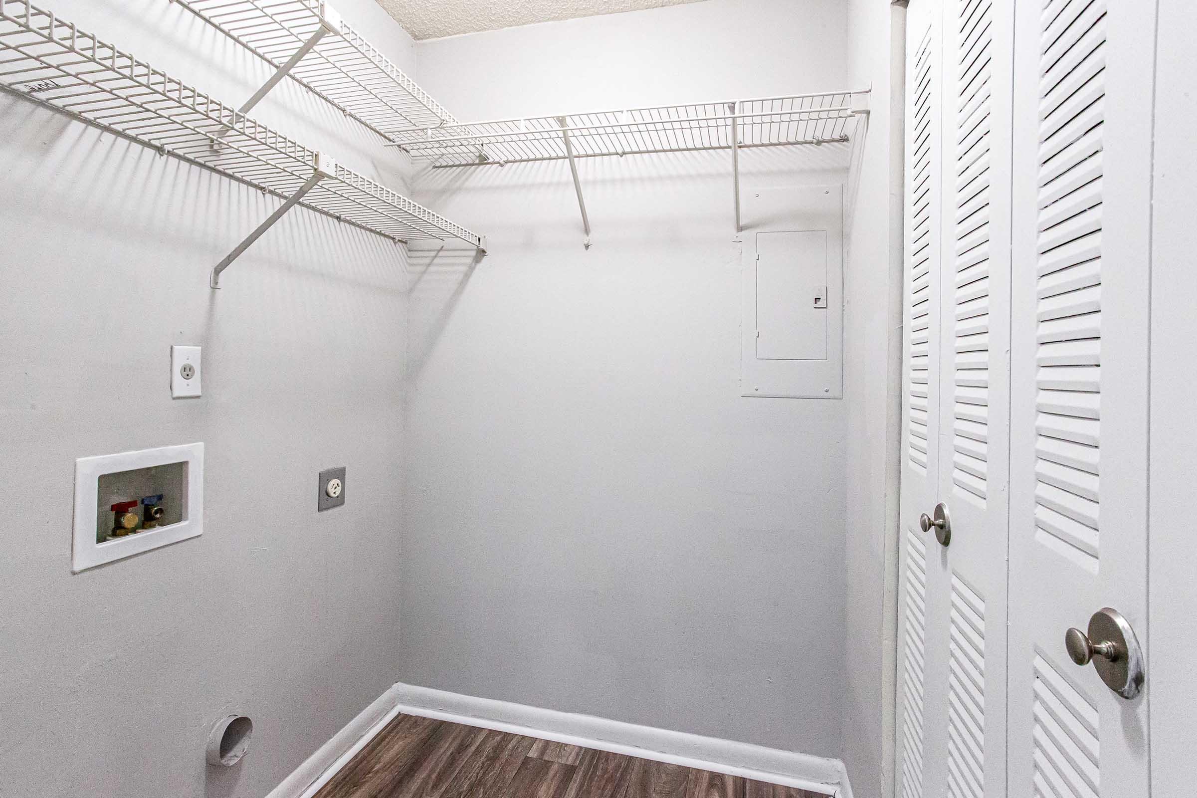 An empty closet with light gray walls and a wooden floor. The closet features wire shelving along the back and side walls, and doors with vertical slats. There’s an electrical panel on the wall and a small outlet visible. The space is tidy and uncluttered, suitable for storage.