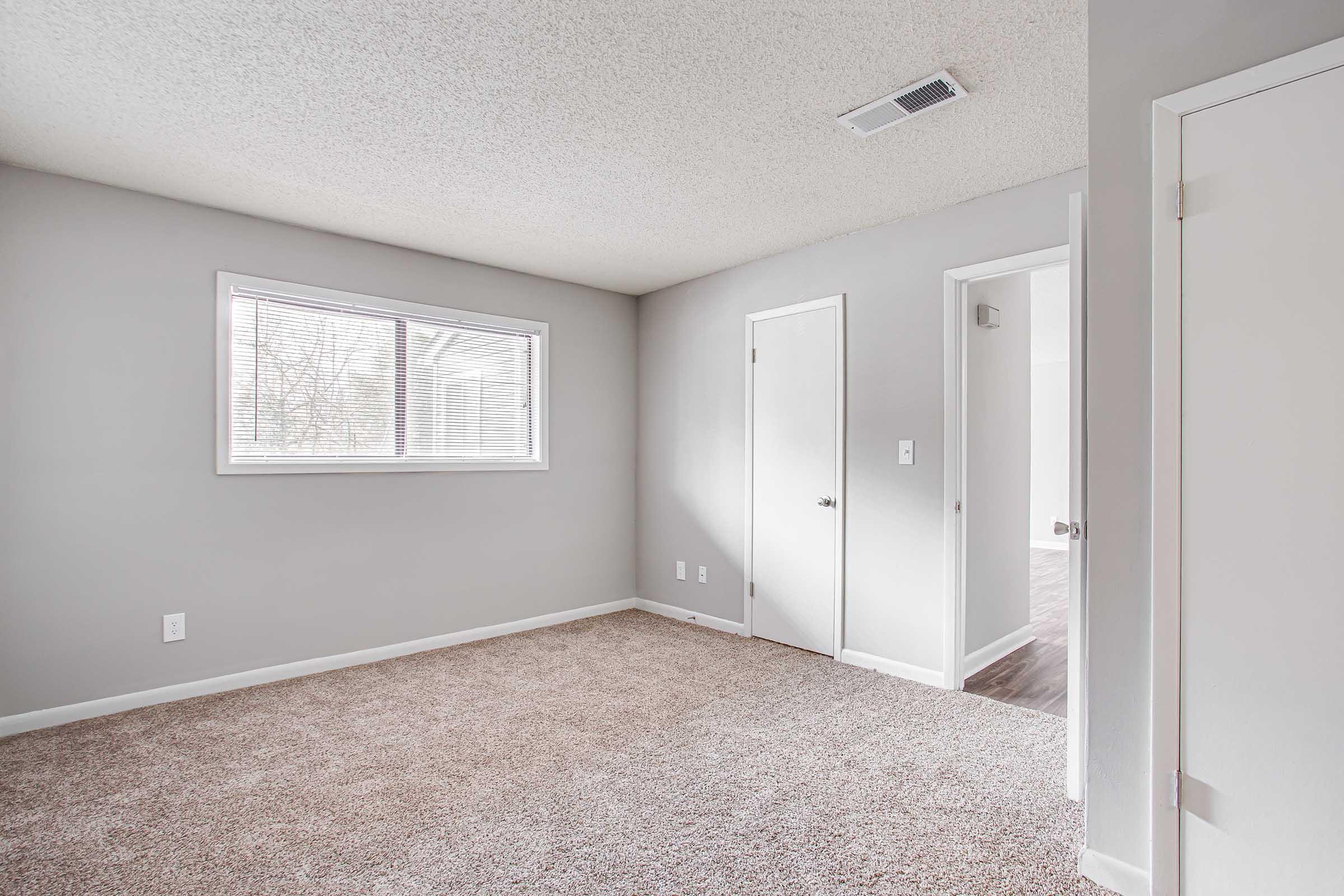 A spacious, unfurnished room featuring light gray walls and a textured ceiling. There is a large window with white blinds, allowing natural light to enter, and a door leading to another room. The floor is covered in beige carpet, creating a cozy atmosphere. The overall look is clean and minimalist.
