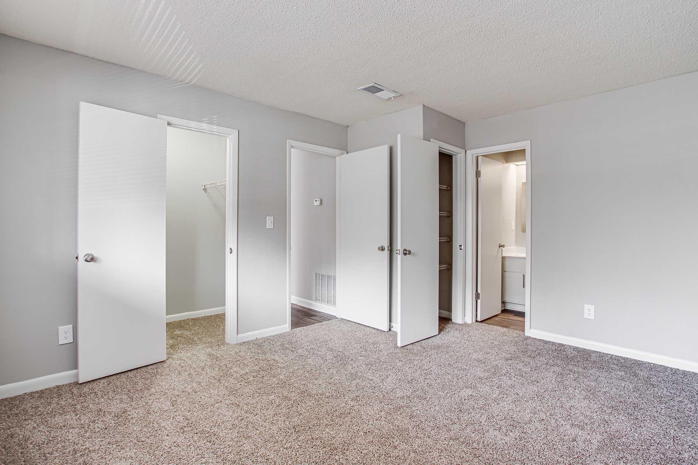 An interior view of a room featuring light gray walls and carpeted flooring. There are multiple doors leading to other spaces, including a closet and bathroom, creating an open and spacious layout. Natural light is coming through the doors, highlighting the neutral decor.