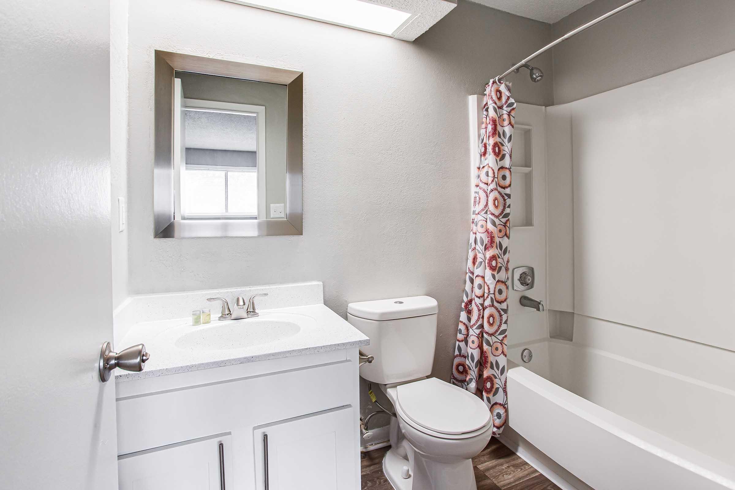 A modern bathroom featuring a white vanity with a sink, a toilet, and a bathtub with a shower curtain displaying a colorful circular pattern. A large mirror hangs above the sink, and there is natural light coming from a window in the background. The walls are a light gray, and the flooring resembles wood.