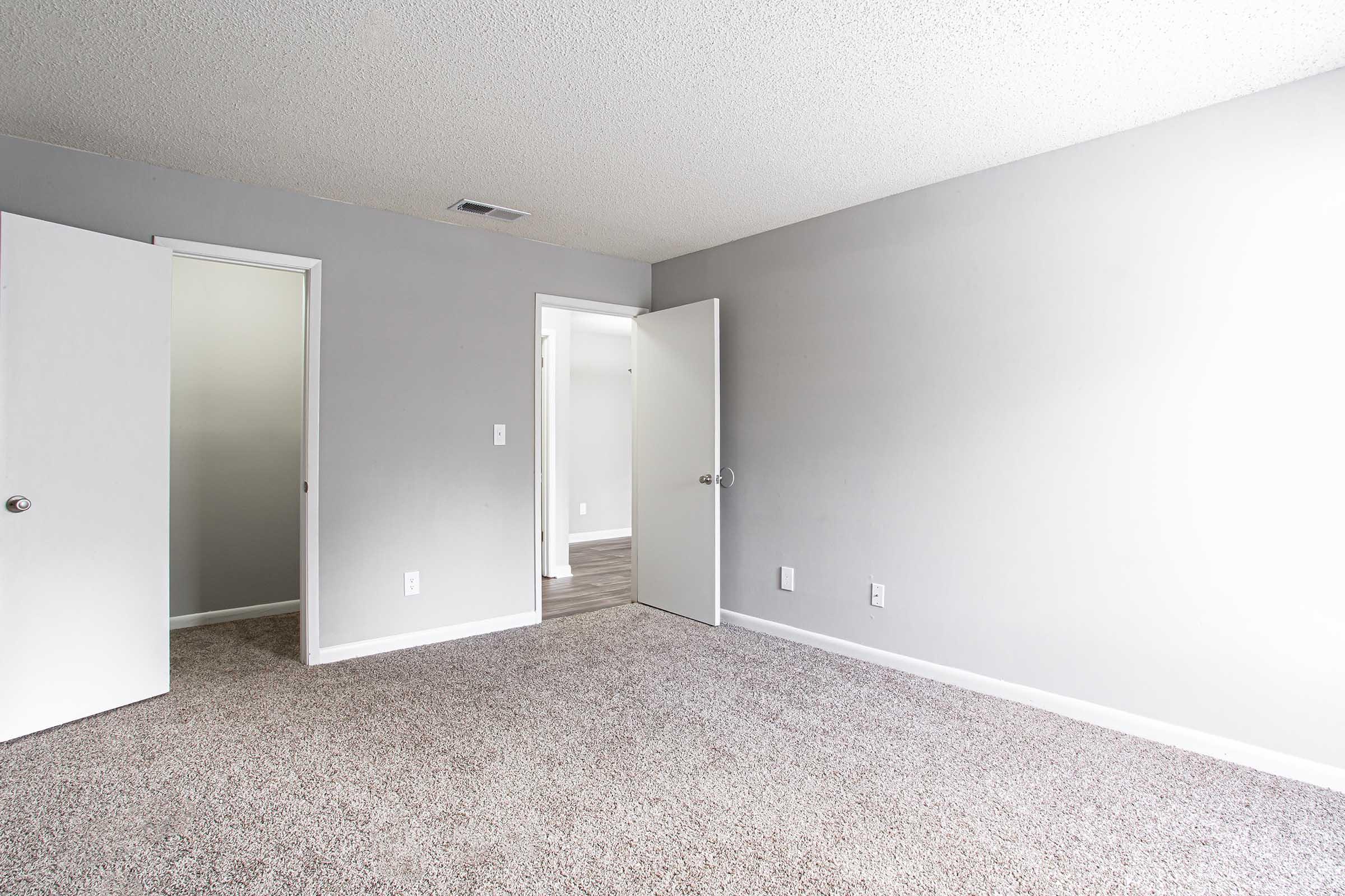 A spacious, empty room with beige carpet and light gray walls. Two open white doors are visible on the left, leading to adjacent areas. The room has good natural light, creating a bright and airy atmosphere, with no furniture or decorations present.