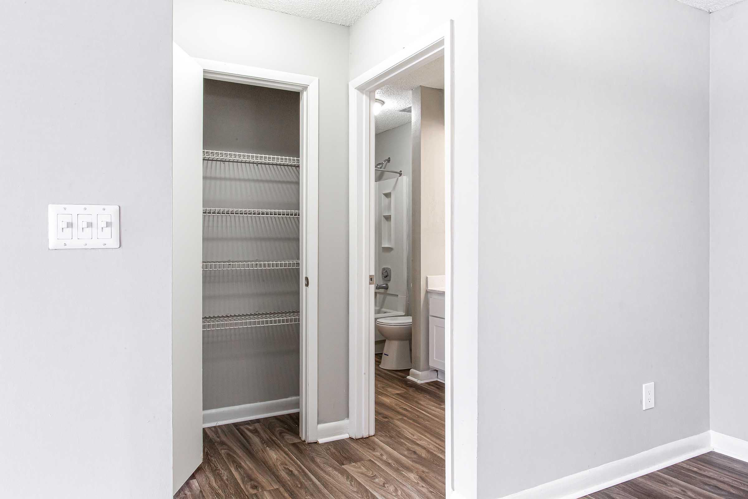 View of a small room with two doorways: one leads to a bathroom with a toilet and sink, and the other opens into a closet with wire shelving. The walls are painted light gray, and the floor has a wood-like finish. Bright, natural light illuminates the space.