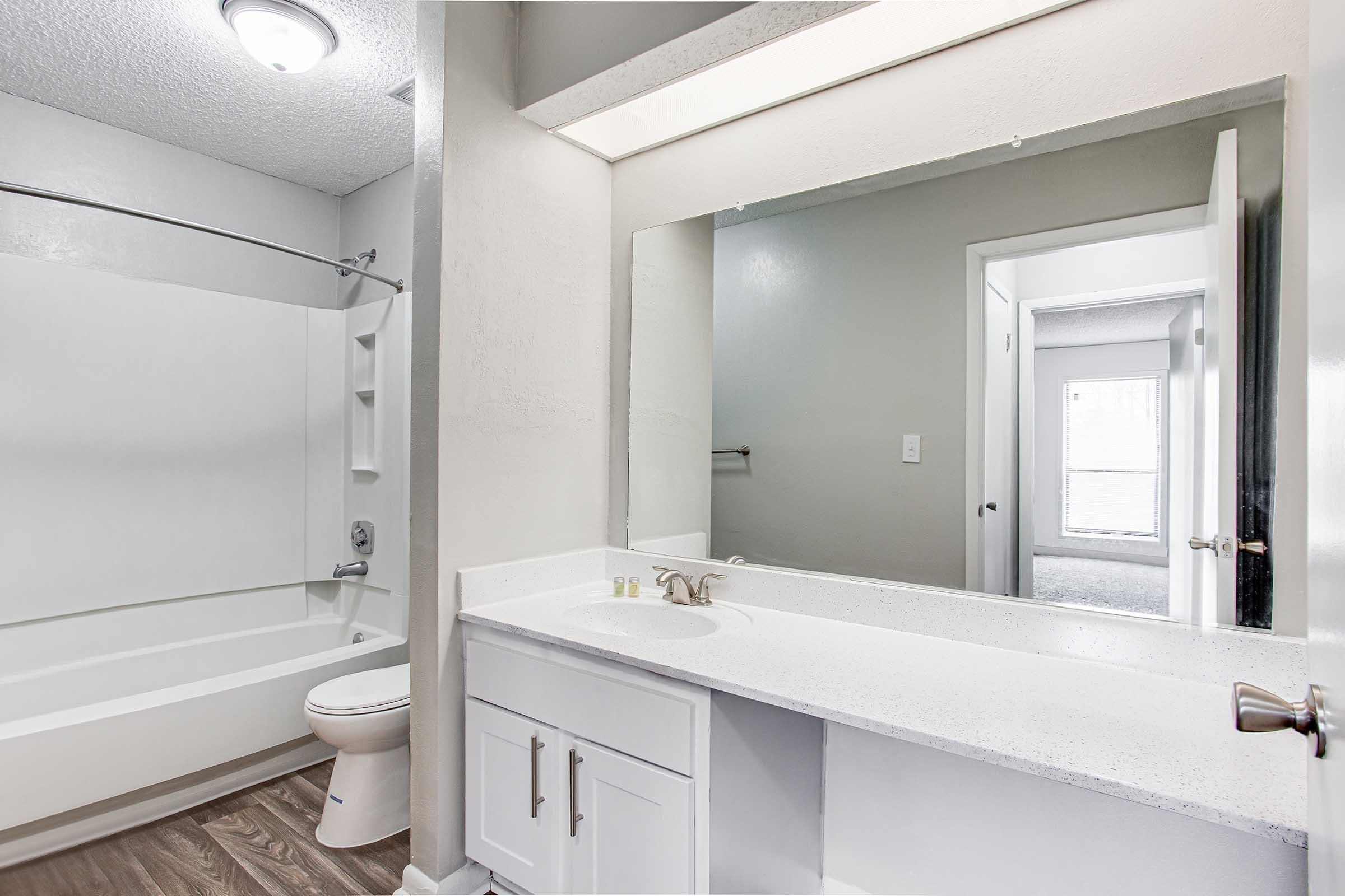 A modern bathroom featuring a white bathtub and shower combination, a sleek white vanity with a single sink, and a large mirror above the vanity. The walls are painted in neutral colors, and there is a light fixture on the ceiling. A window can be seen in the background, allowing natural light to enter.