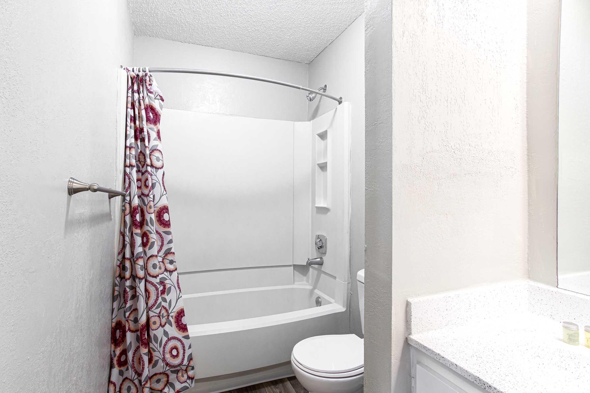 A clean bathroom featuring a shower/tub combo with a floral shower curtain, a modern toilet, and a vanity with a light-colored countertop. The walls are painted in neutral tones, and the space is well-lit.