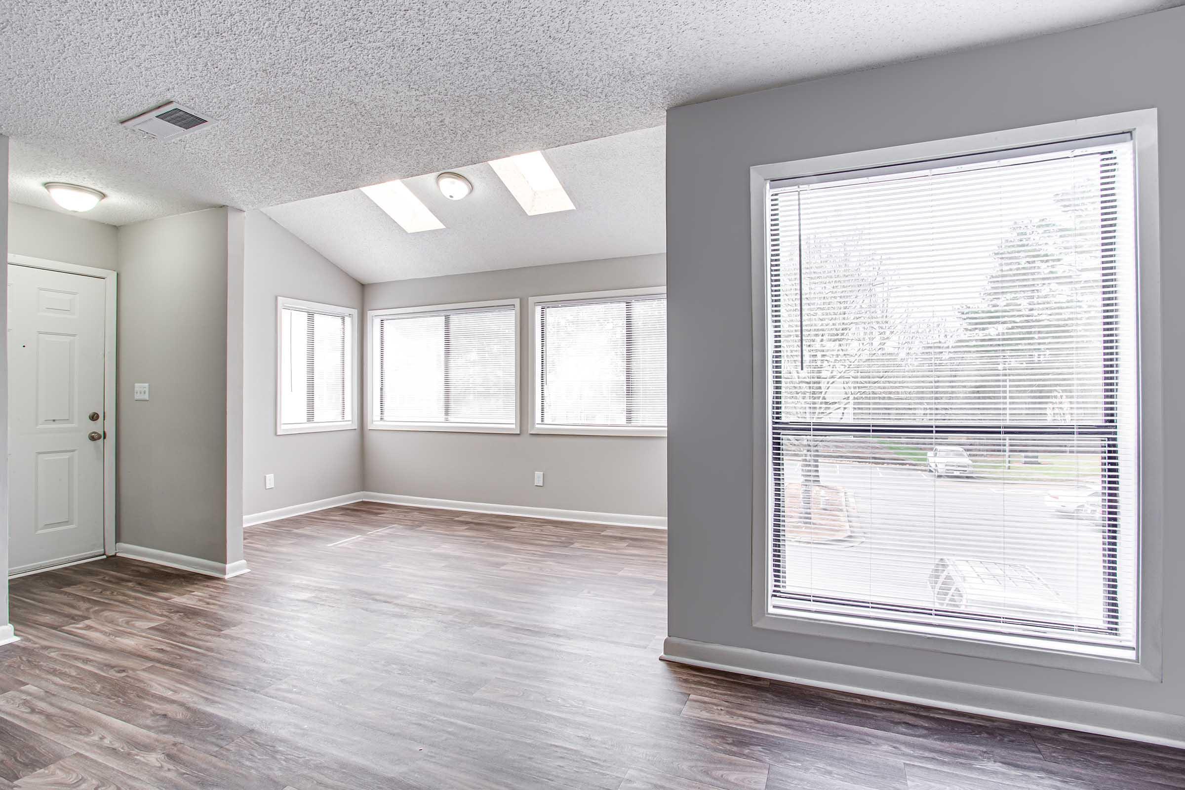 Bright and spacious living area with large windows allowing natural light, light gray walls, and wooden floors. A front door is visible on the left, and two skylights add to the brightness. The corner provides a view outside, enhancing the open feel of the space.