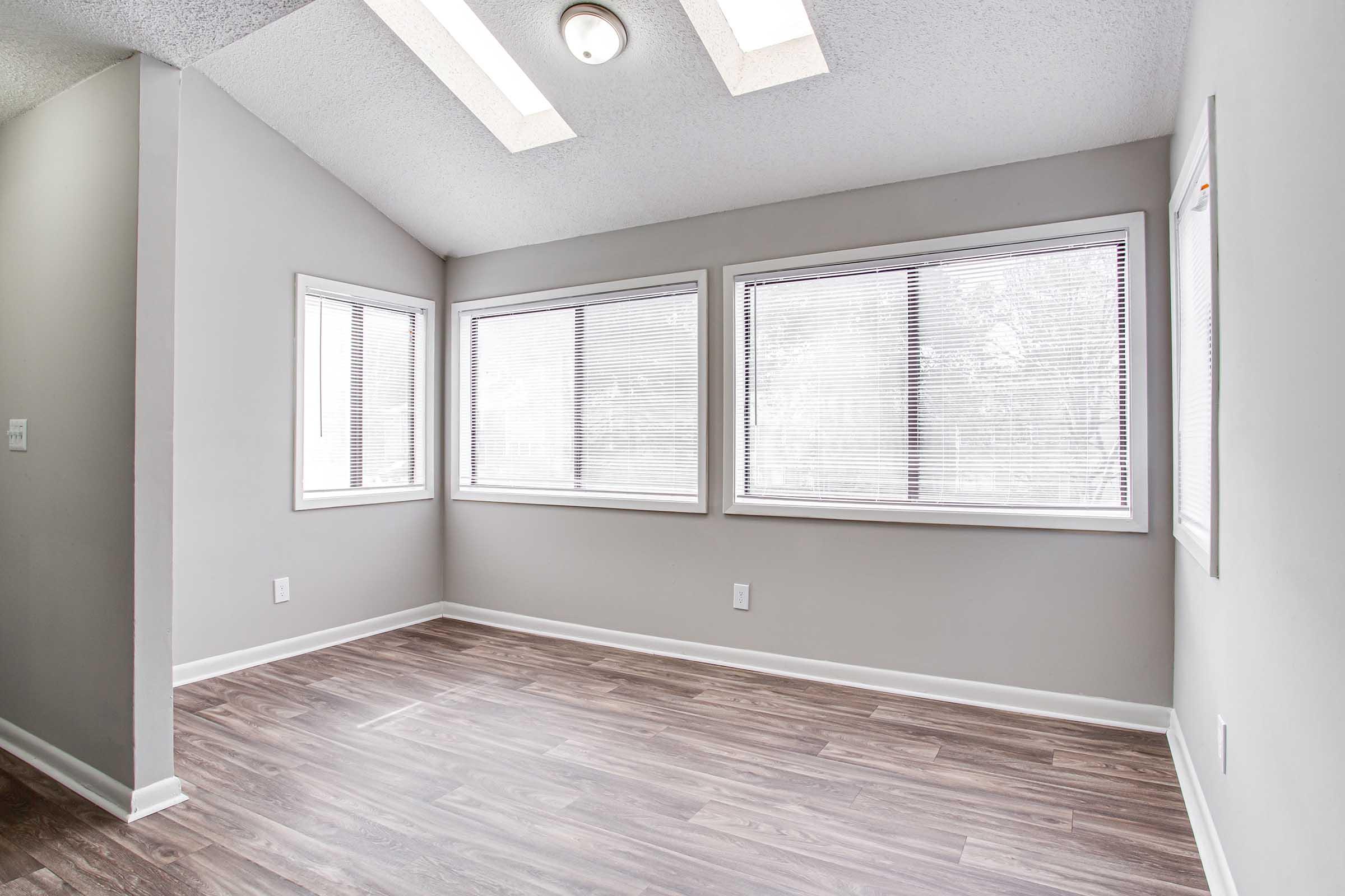 A well-lit, empty room with gray walls and wooden flooring. There are three large windows with blinds letting in natural light, and a ceiling light fixture above. The space has a clean, modern aesthetic, making it suitable for various uses.